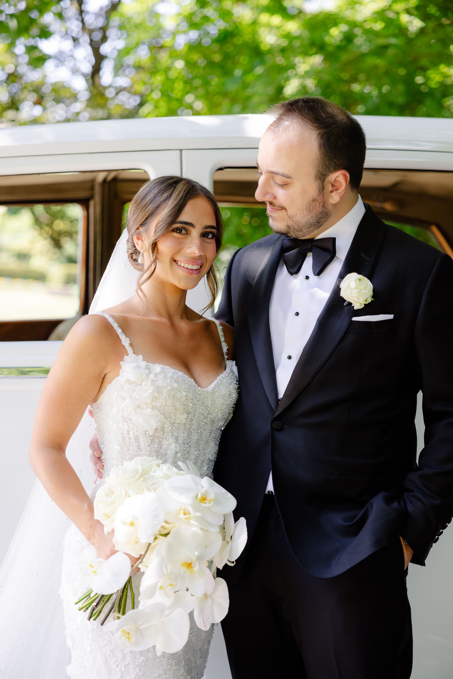 A bride in a white lace gown holds white orchid bouquet while standing with groom in classic tuxedo next to vintage car.