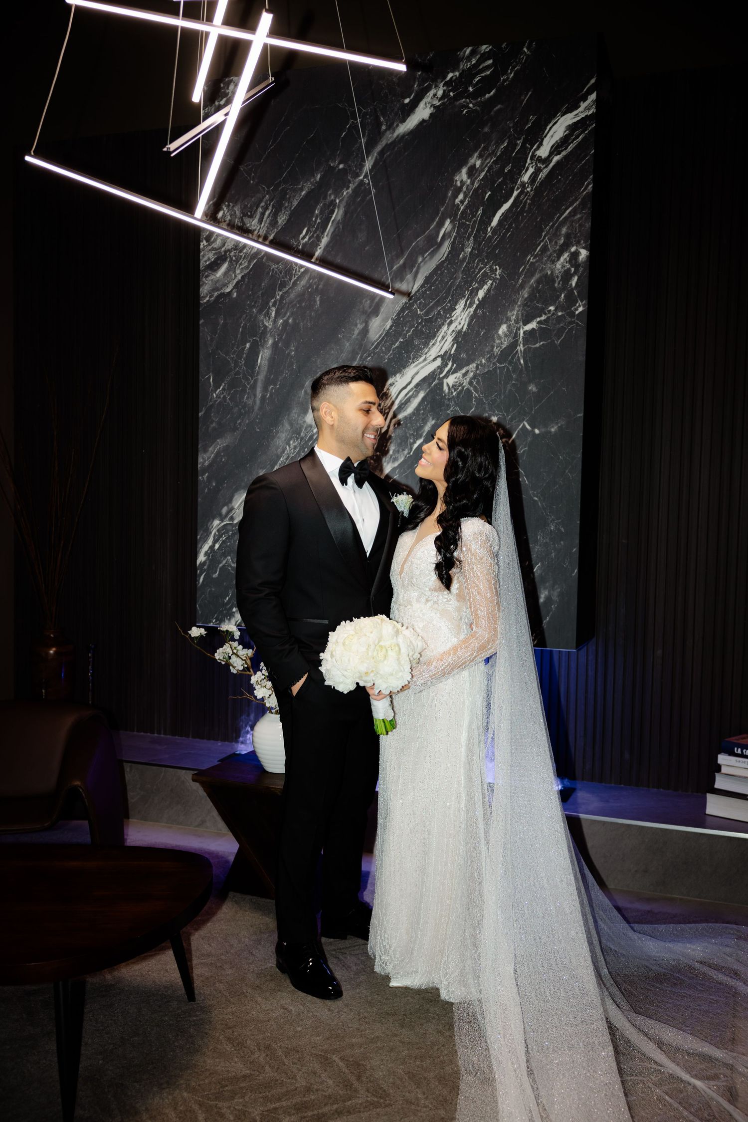 Wedding couple stands under modern lighting with marble wall backdrop and white floral bouquet.