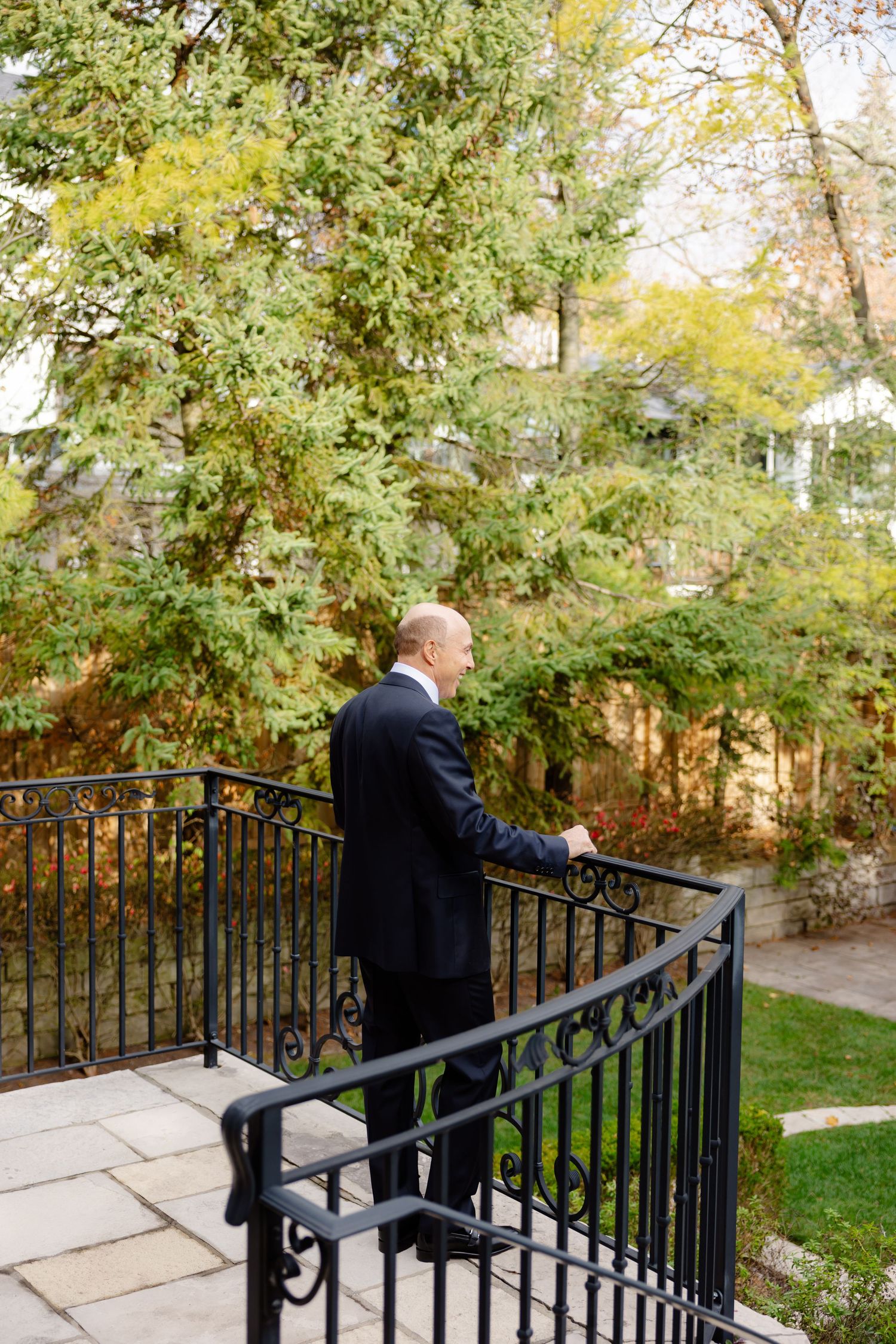Man in dark suit stands on curved outdoor staircase with iron railing surrounded by autumn foliage.