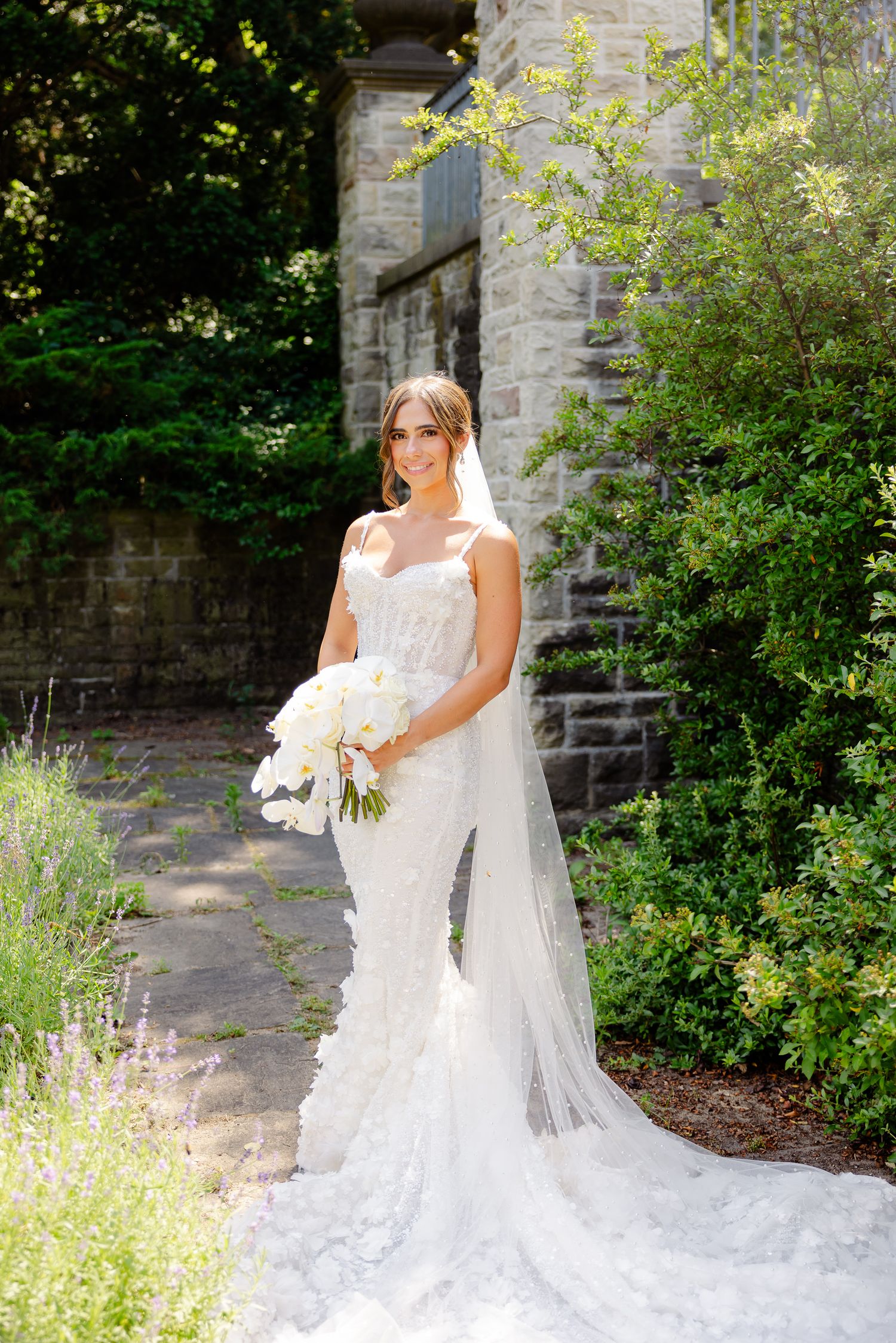 A bride in a flowing white lace wedding dress poses in a garden setting with stone walls and greenery.