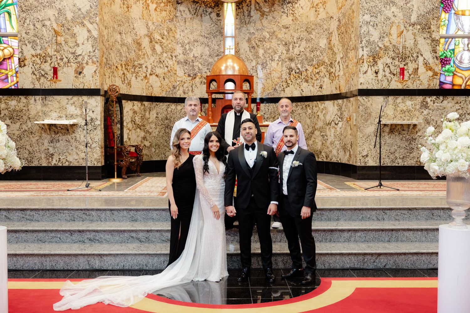 Wedding party poses on church altar steps with ornate cross and stained glass windows in background.