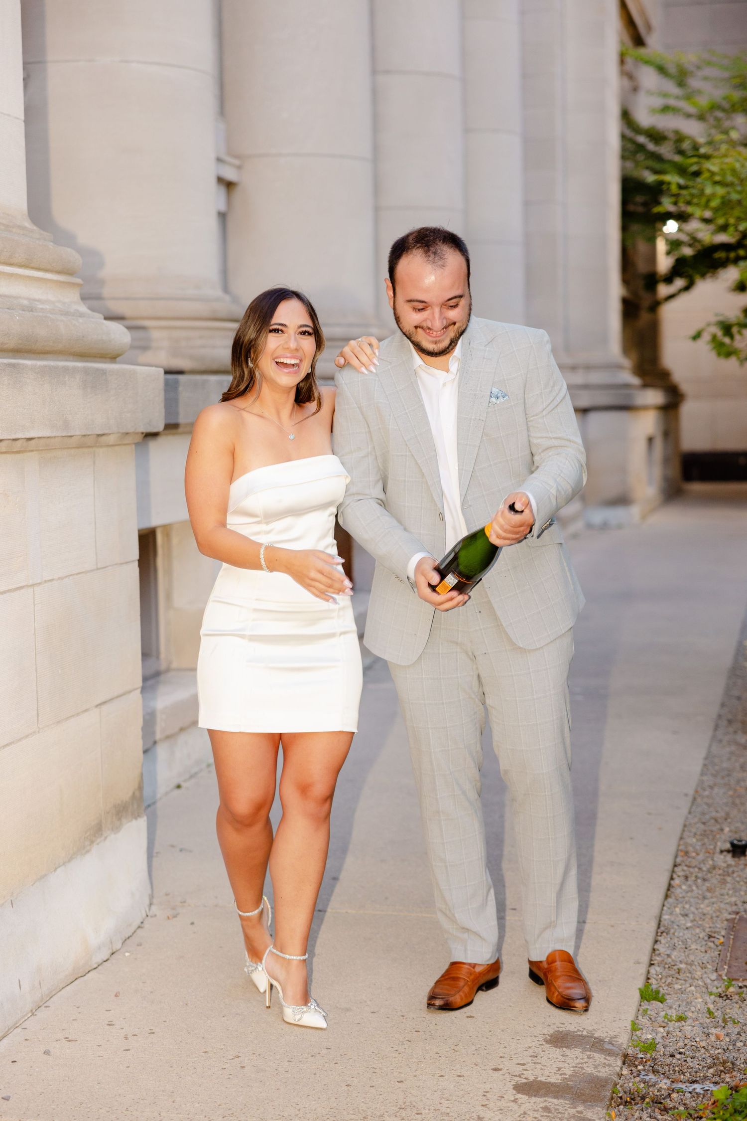 A celebratory couple laughs while holding champagne against a columned building backdrop.