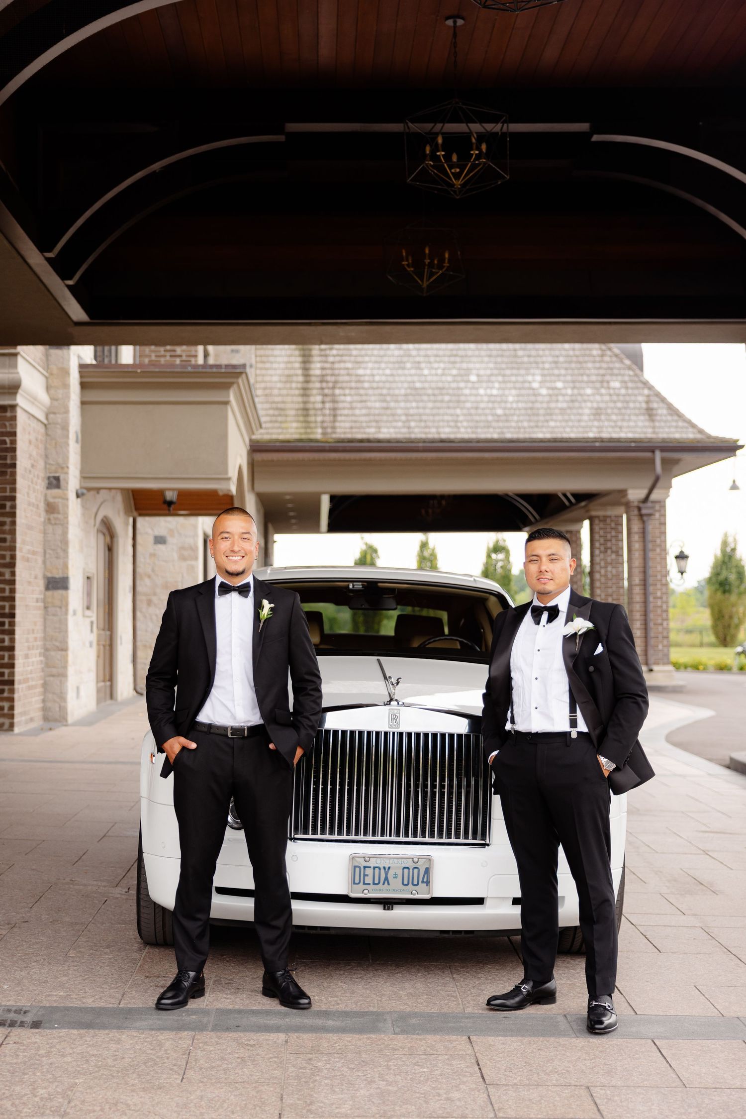 Low angle view of formal tuxedo shoes and pant legs standing in front of a luxury white car at an outdoor venue.