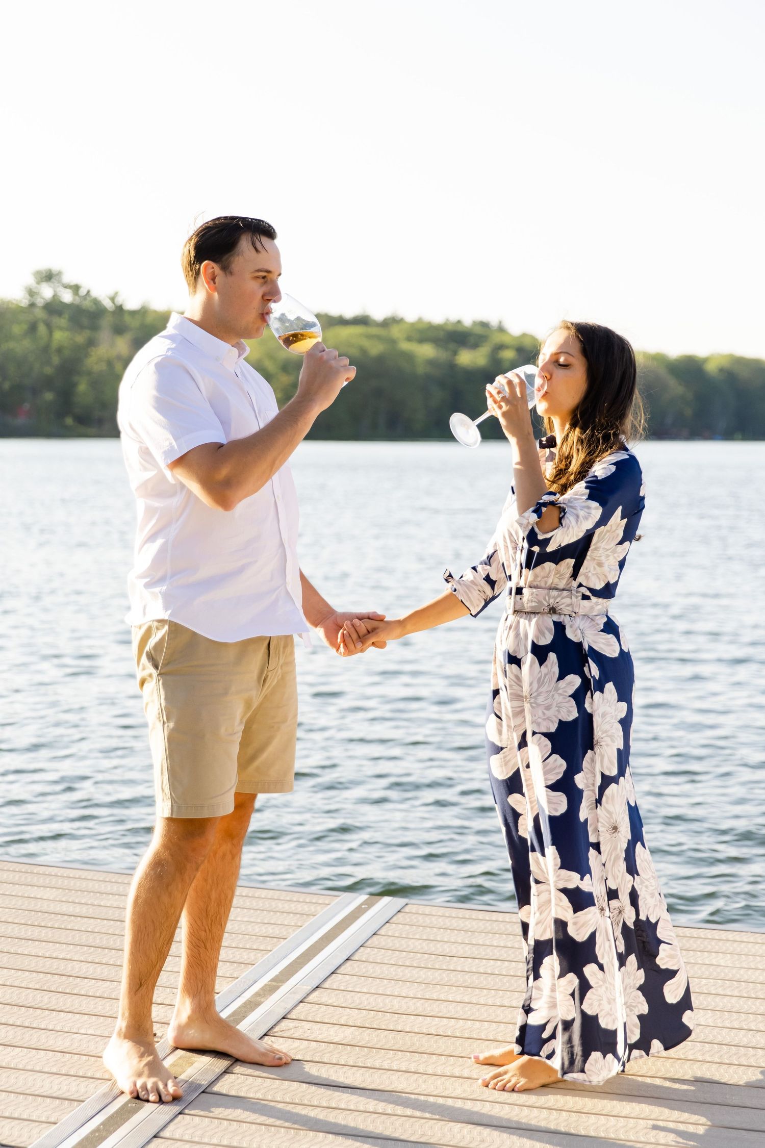 Playful couple sharing drinks and holding hands on wooden dock by the lake.