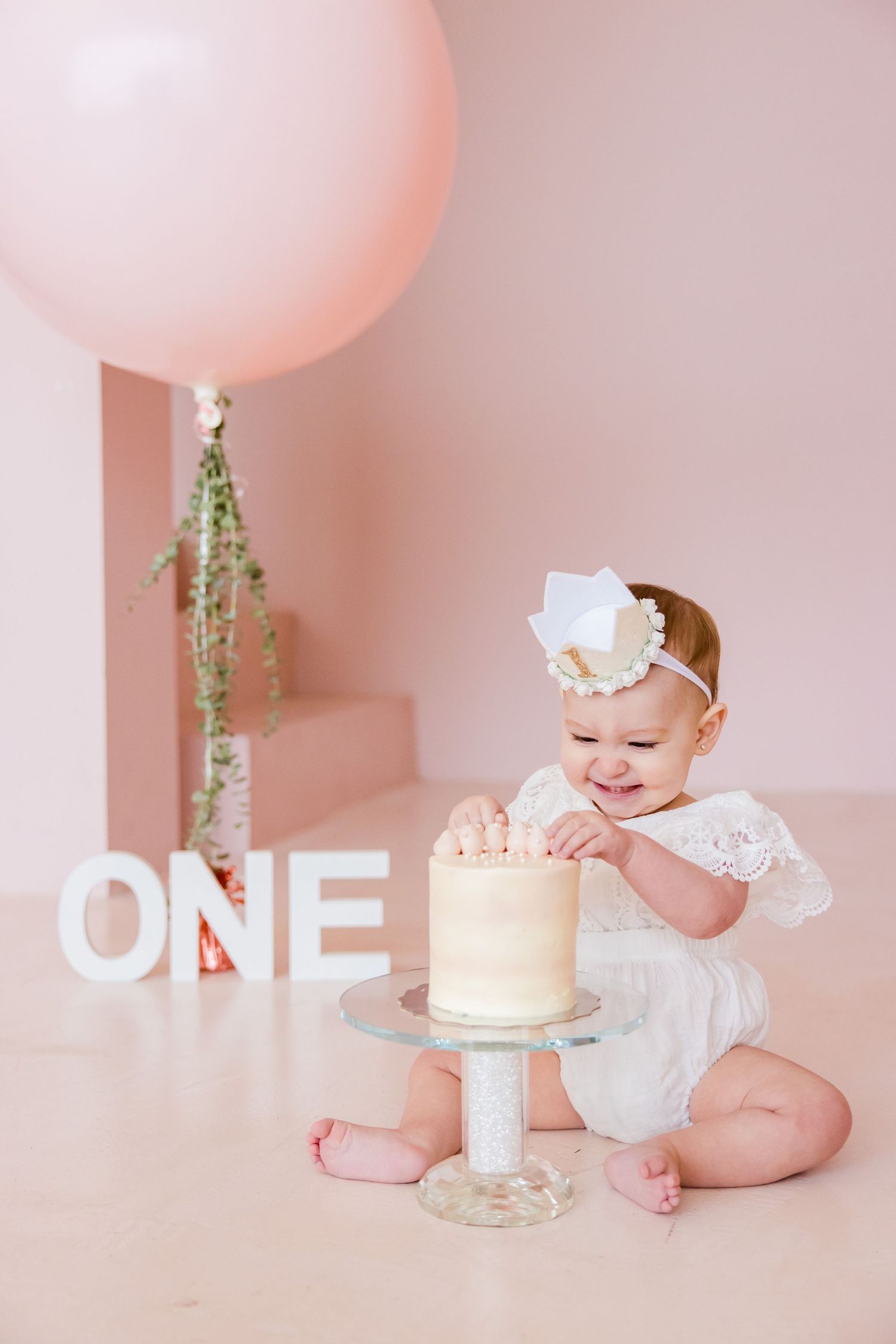 First birthday photo of baby reaching for white cake with large pink balloon and ONE letters.