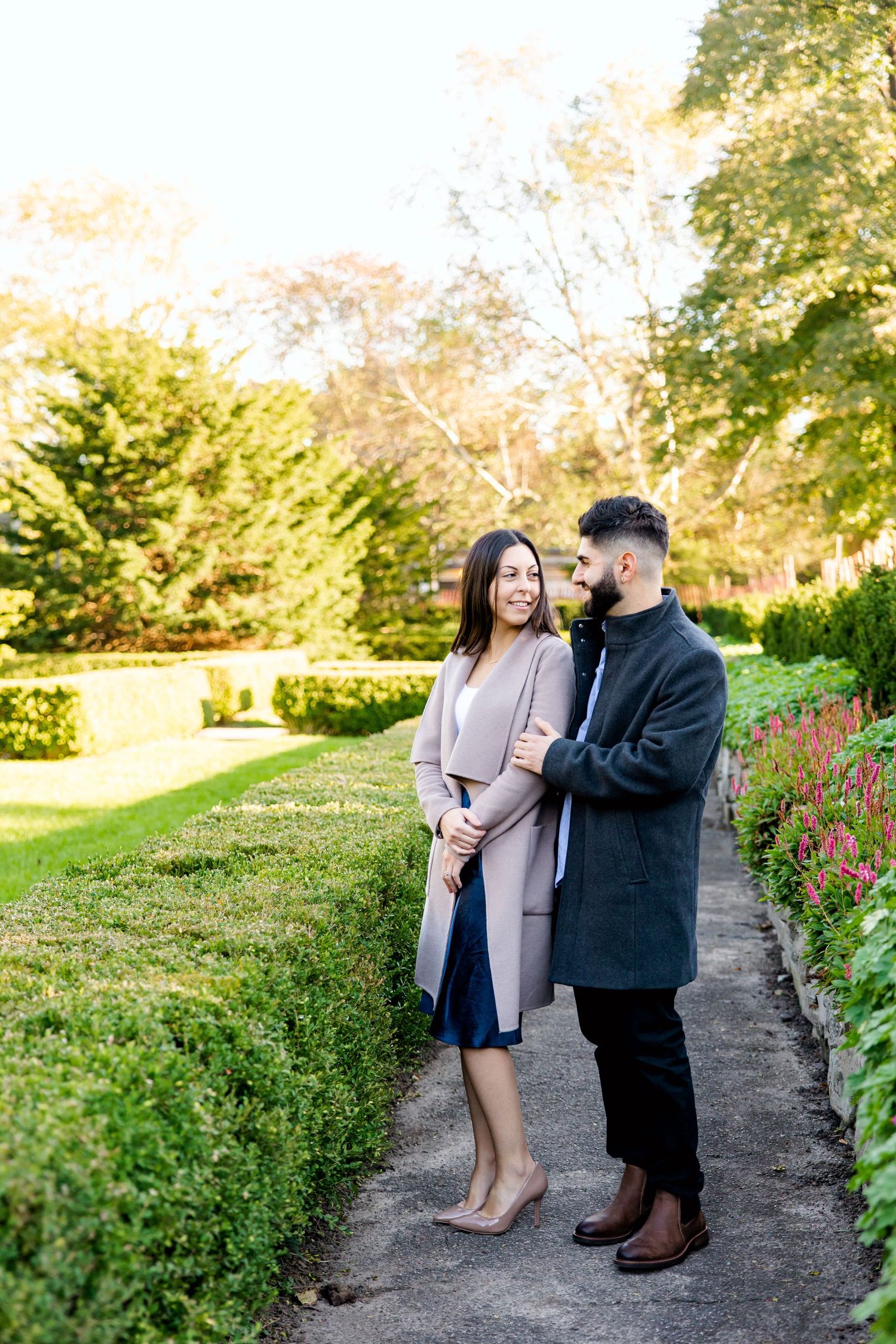 A couple poses together on a garden path surrounded by blooming flowers and manicured hedges.