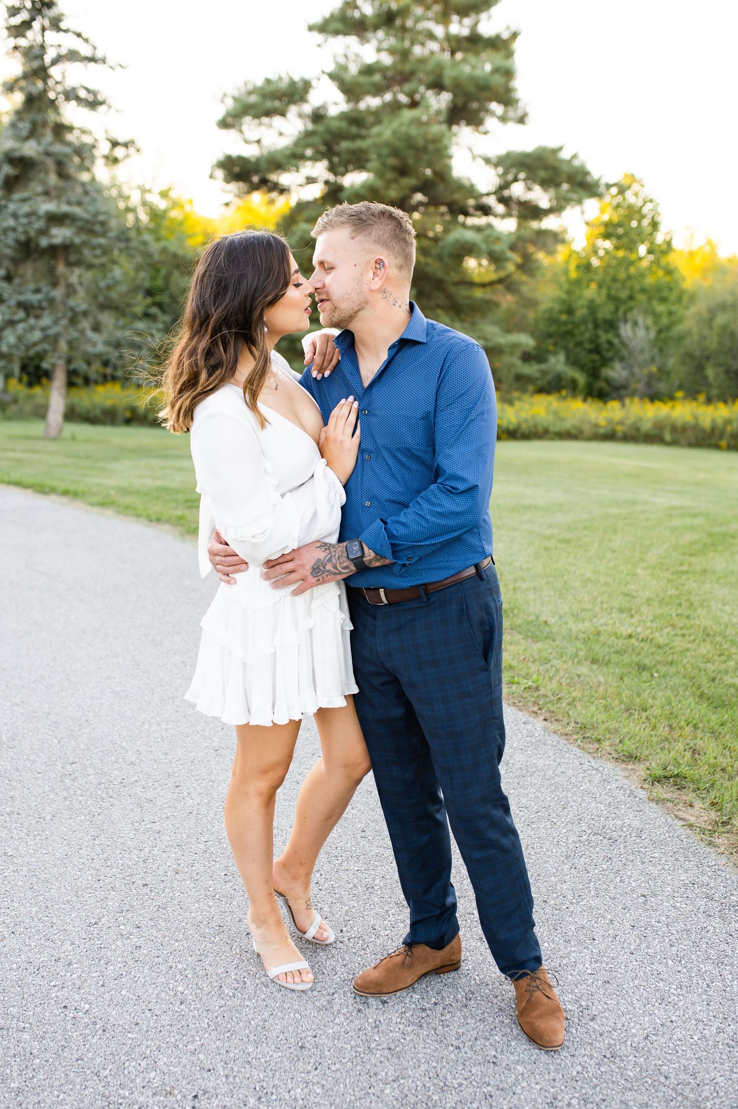 Couple sharing intimate moment on path with green trees and lawn in background.