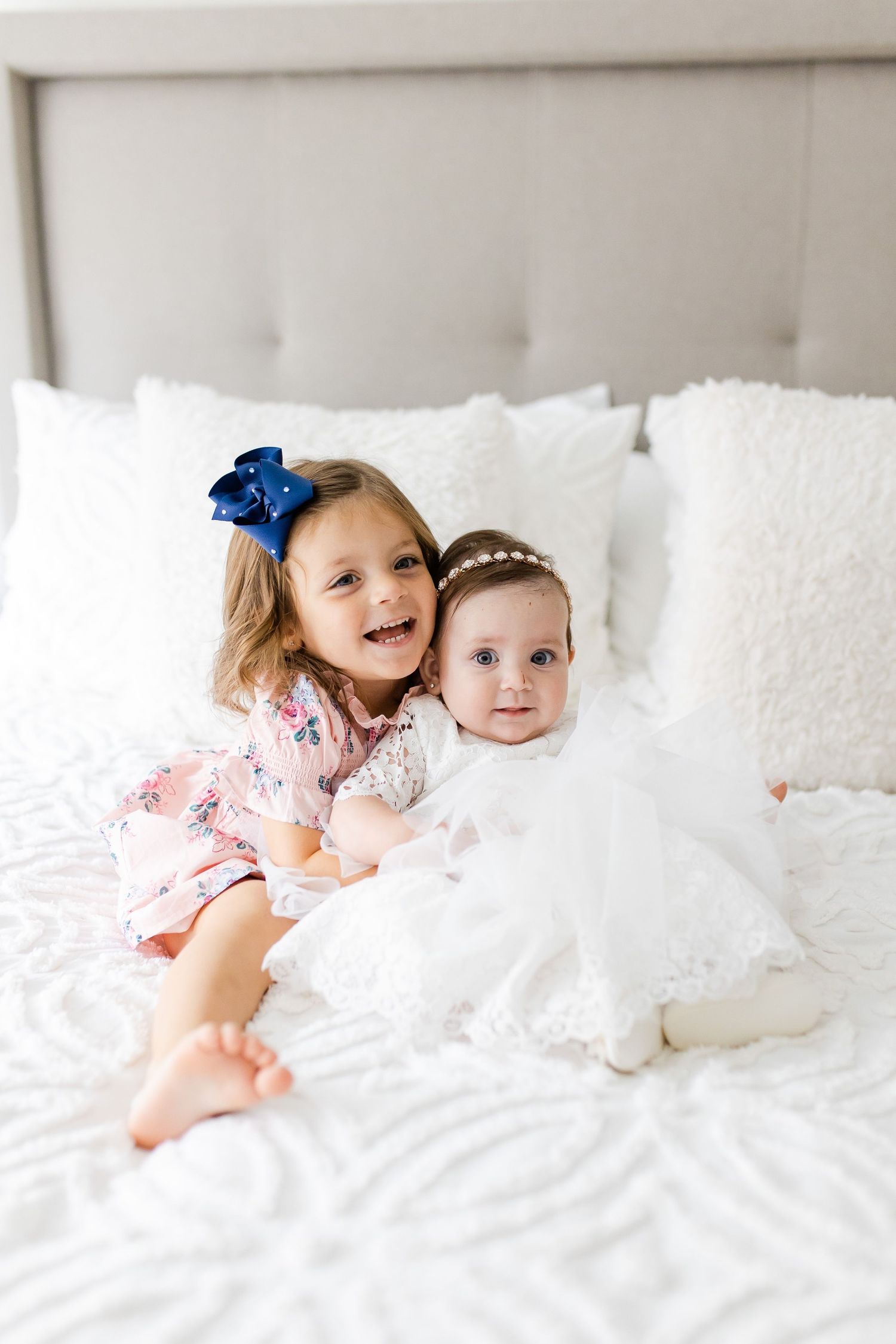 Two young children in white and pink dresses sitting together on white bedding.