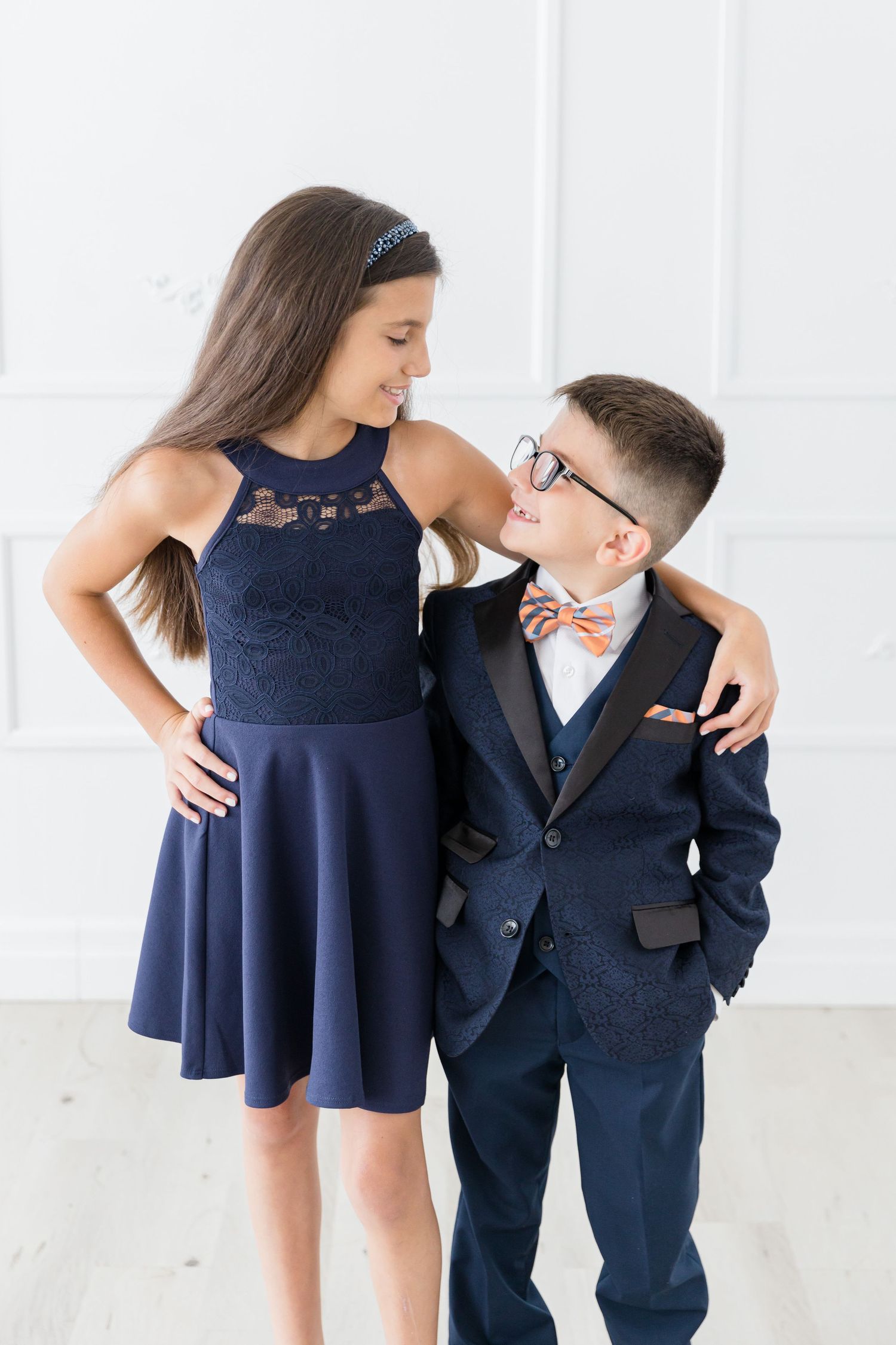 Two people in formal wear share an affectionate moment against white wall background.