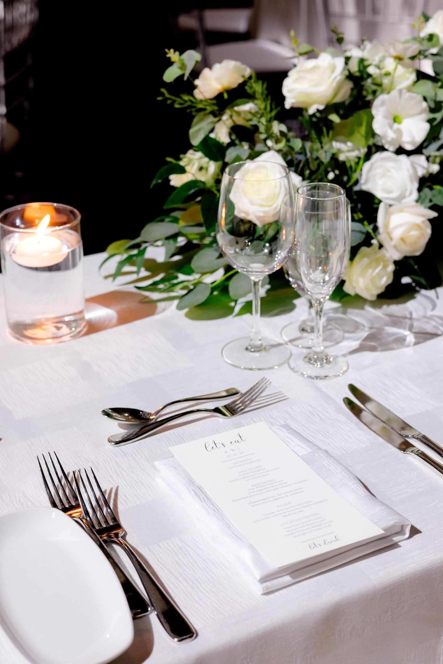 Elegant white wedding table setting with crystal glasses, white roses, and menu card on crisp white tablecloth.