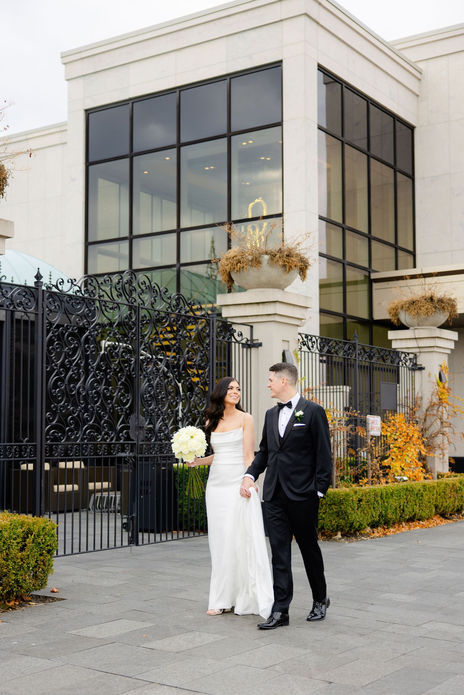 A bride in white gown and groom in tuxedo stand together near decorative gates and glass building facade.