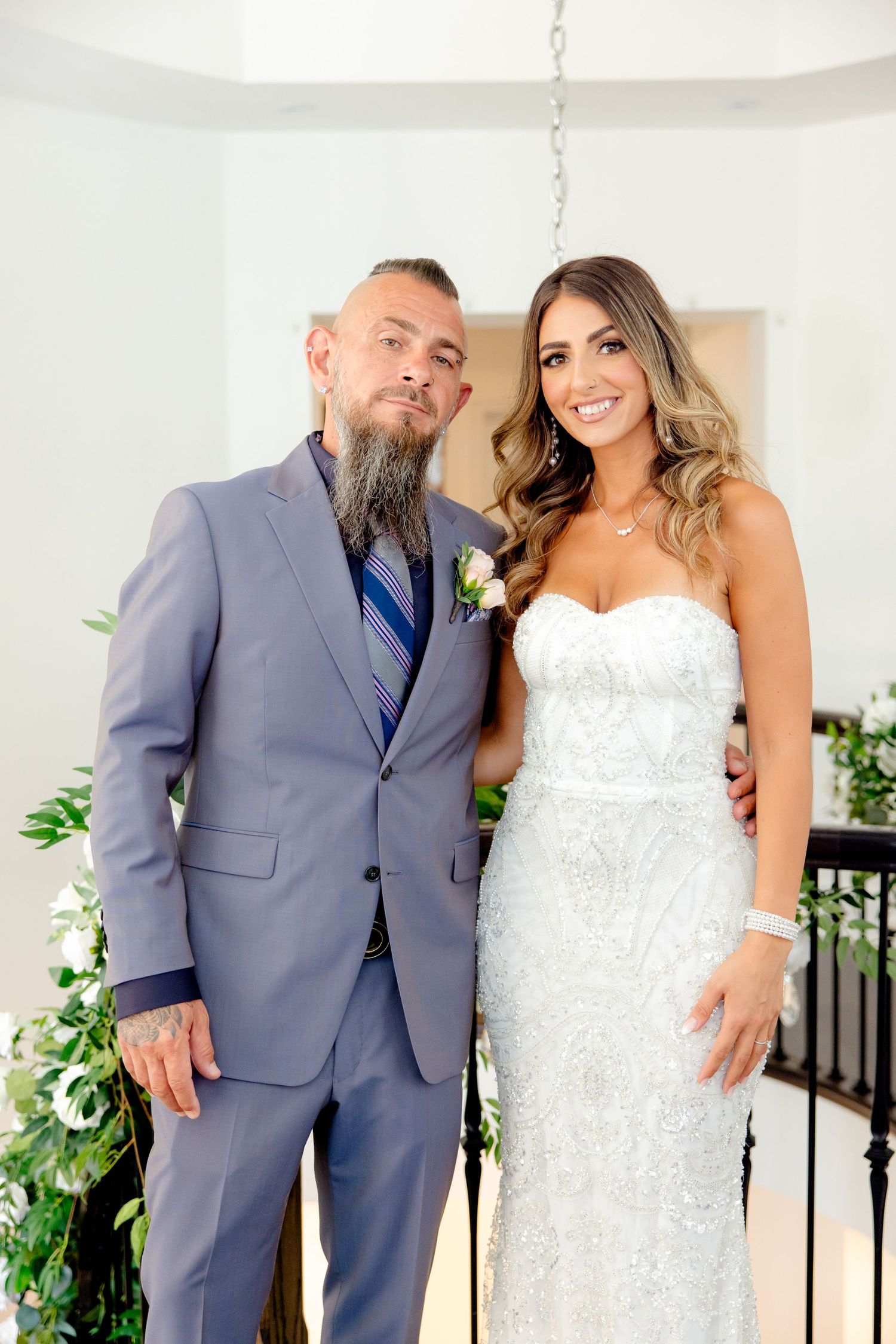 Wedding portrait with couple in formal attire against white wall with greenery.