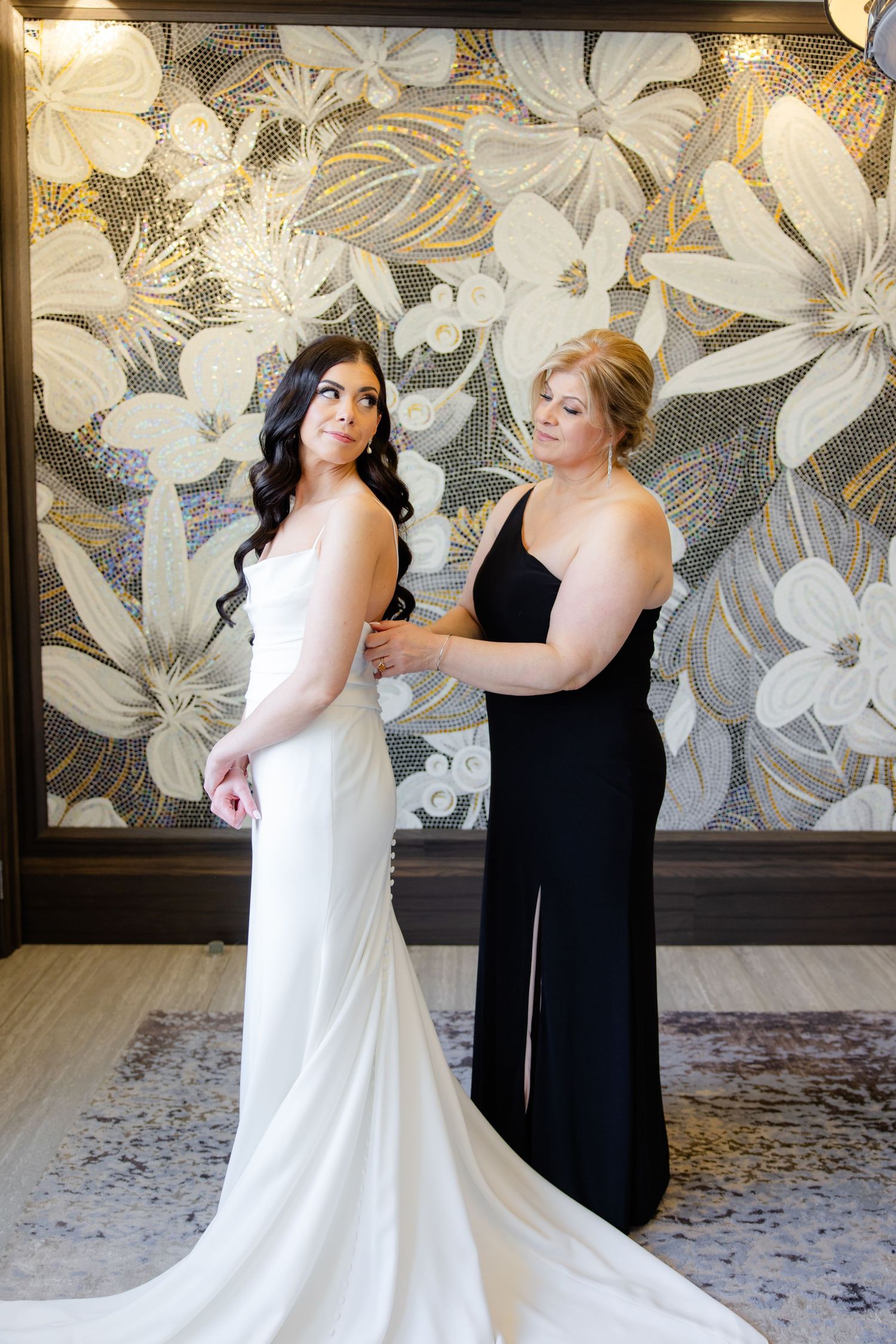 A wedding scene with a white gown being adjusted against a floral wallpaper backdrop.