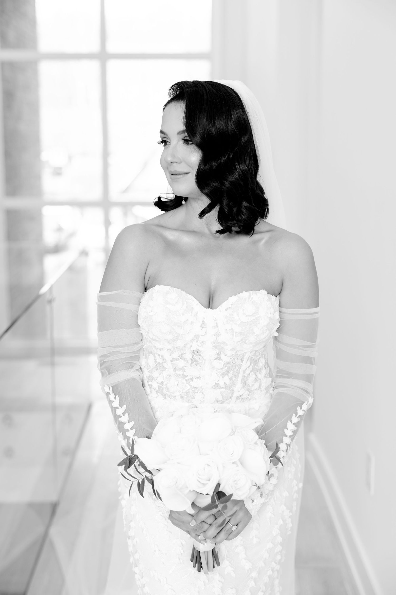 Black and white bridal portrait showing romantic wavy hair and strapless lace gown with sheer sleeves.
