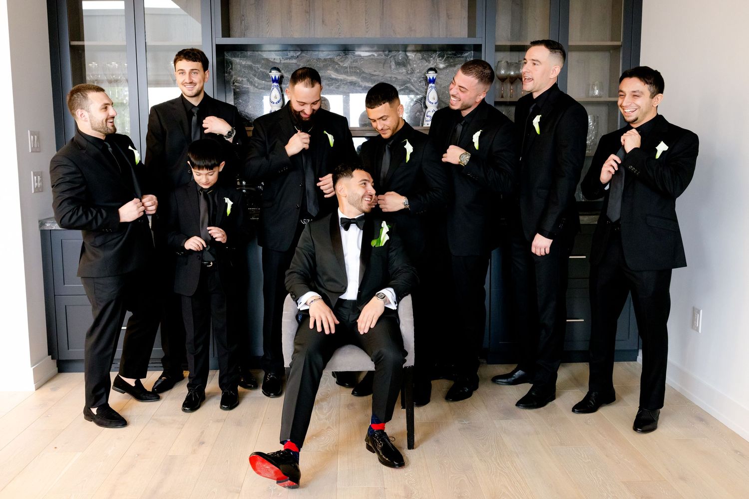 A color photo of groomsmen in black suits gathered around a central seated figure.