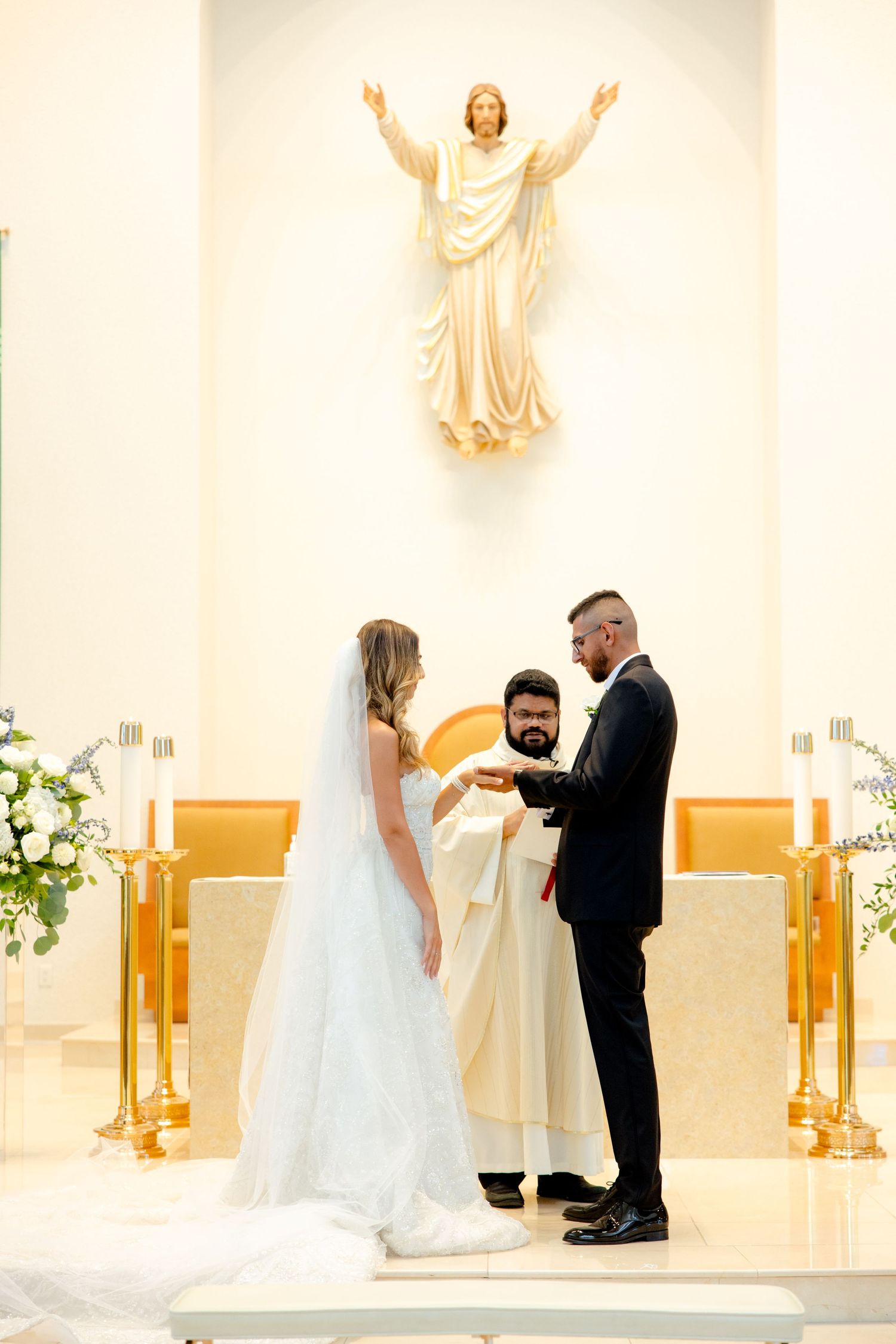 Religious wedding ceremony taking place beneath a decorative Jesus statue in white church interior.