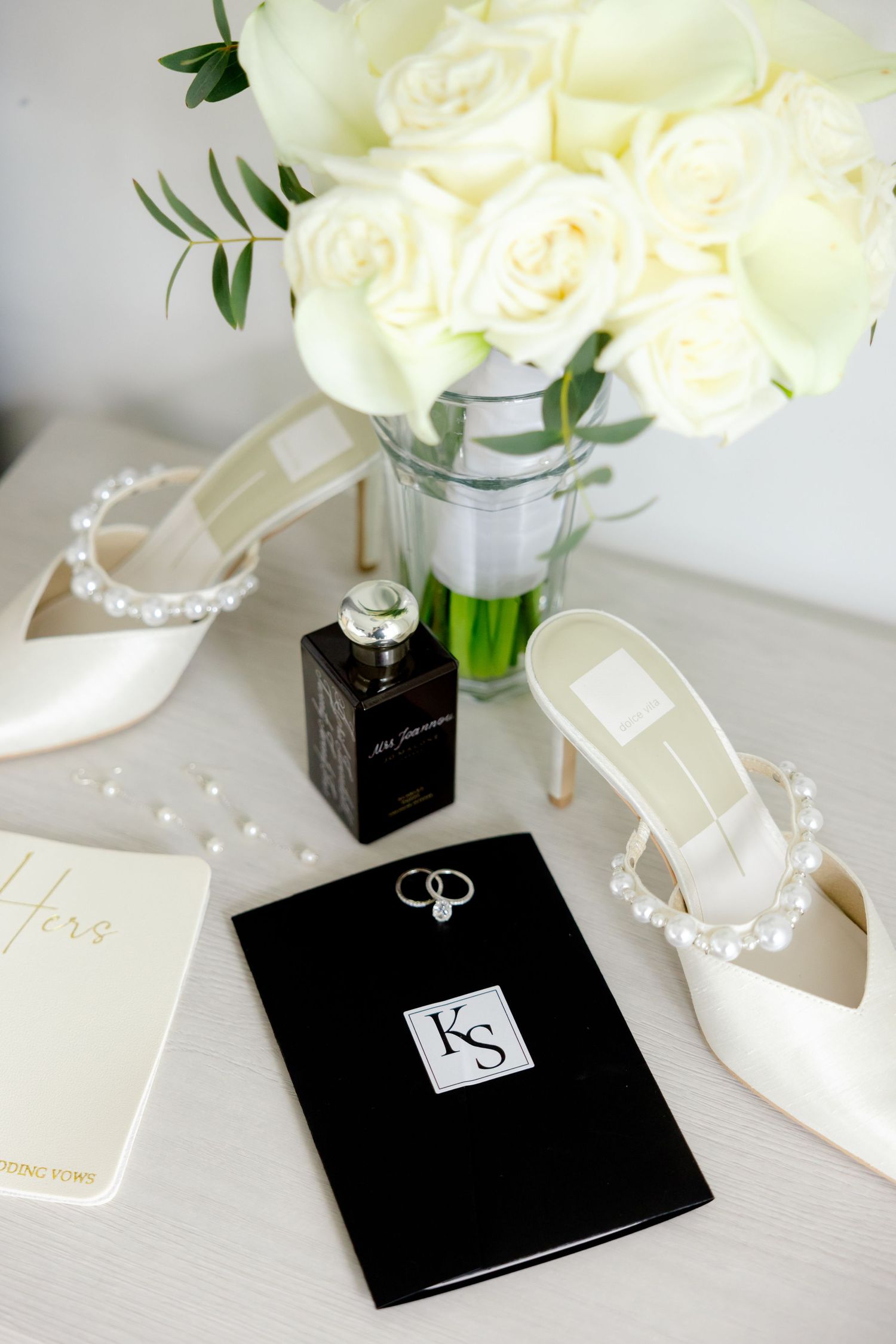 Wedding details flat lay with white roses, pearl shoes, rings and black monogrammed invitation.
