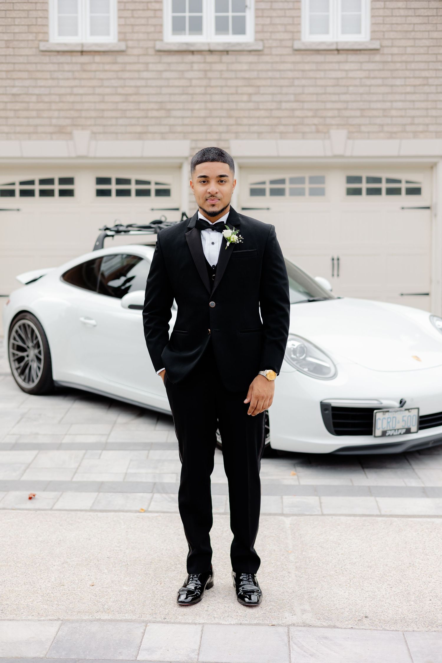 Formal portrait against a white Porsche and brick garage featuring someone in a classic black tuxedo.
