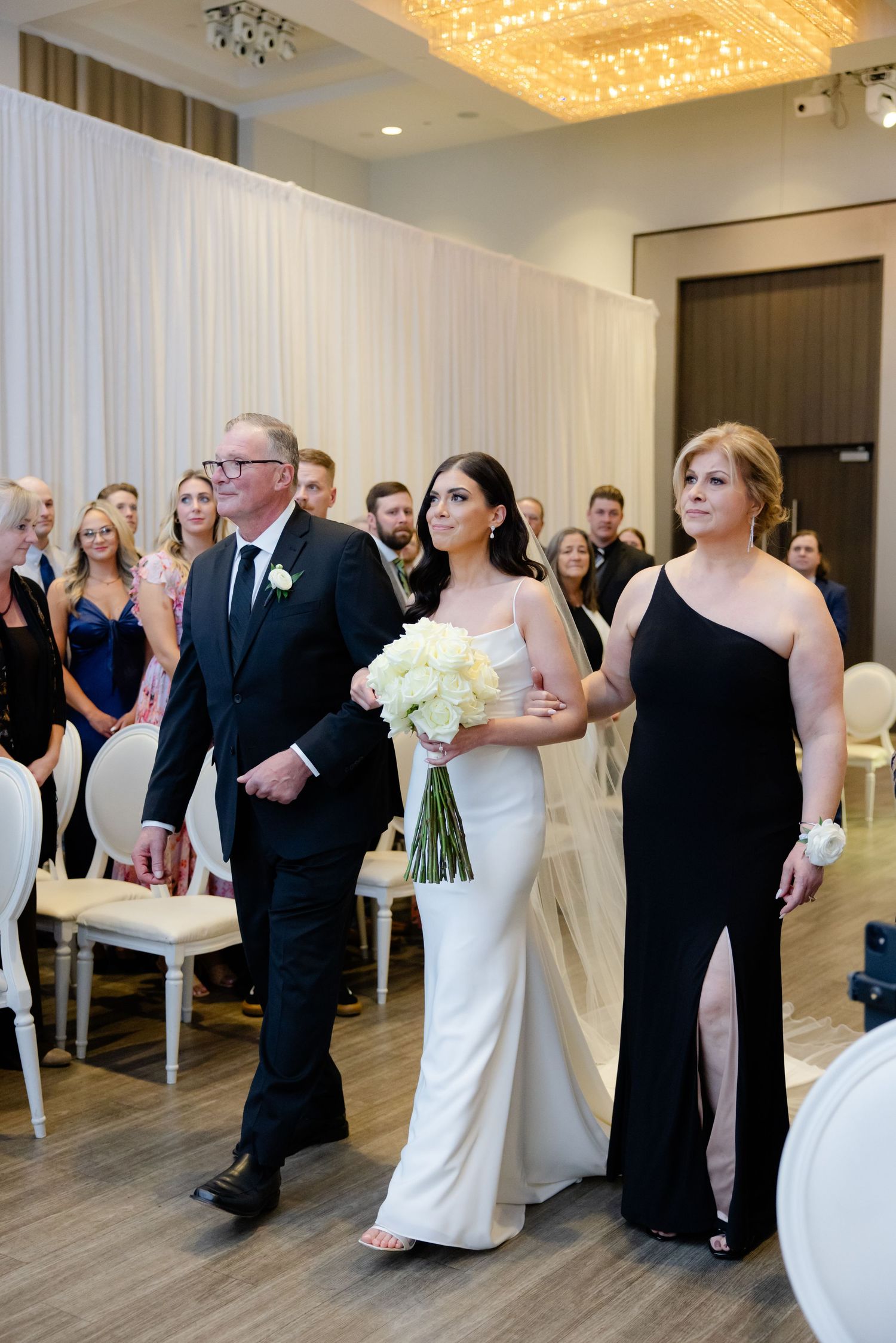 Wedding processional with elegant white and black attire in front of draped white backdrop under crystal chandeliers.