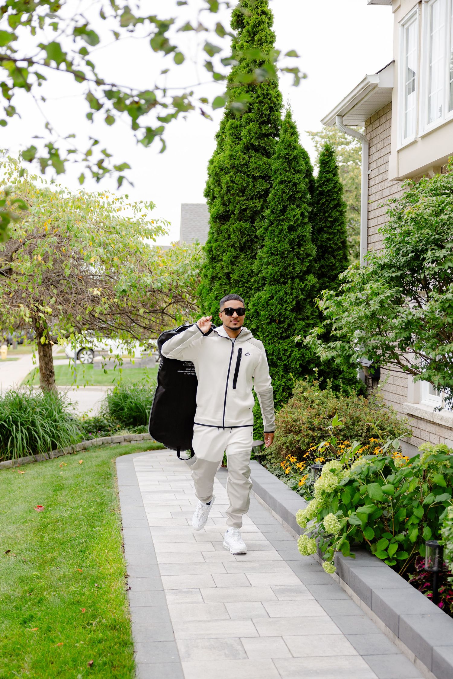 A person in a stylish white tracksuit and sunglasses carries a garment bag along a landscaped path.