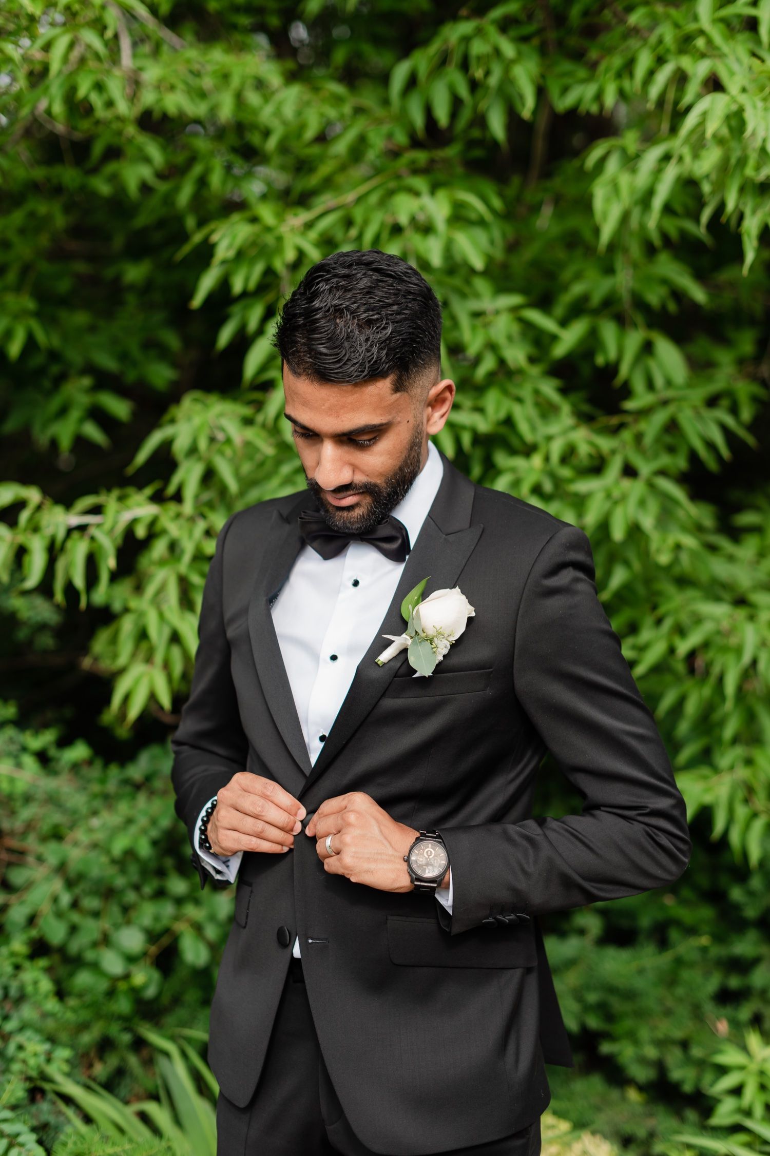 Groom adjusts bowtie wearing classic black tuxedo against green foliage background.