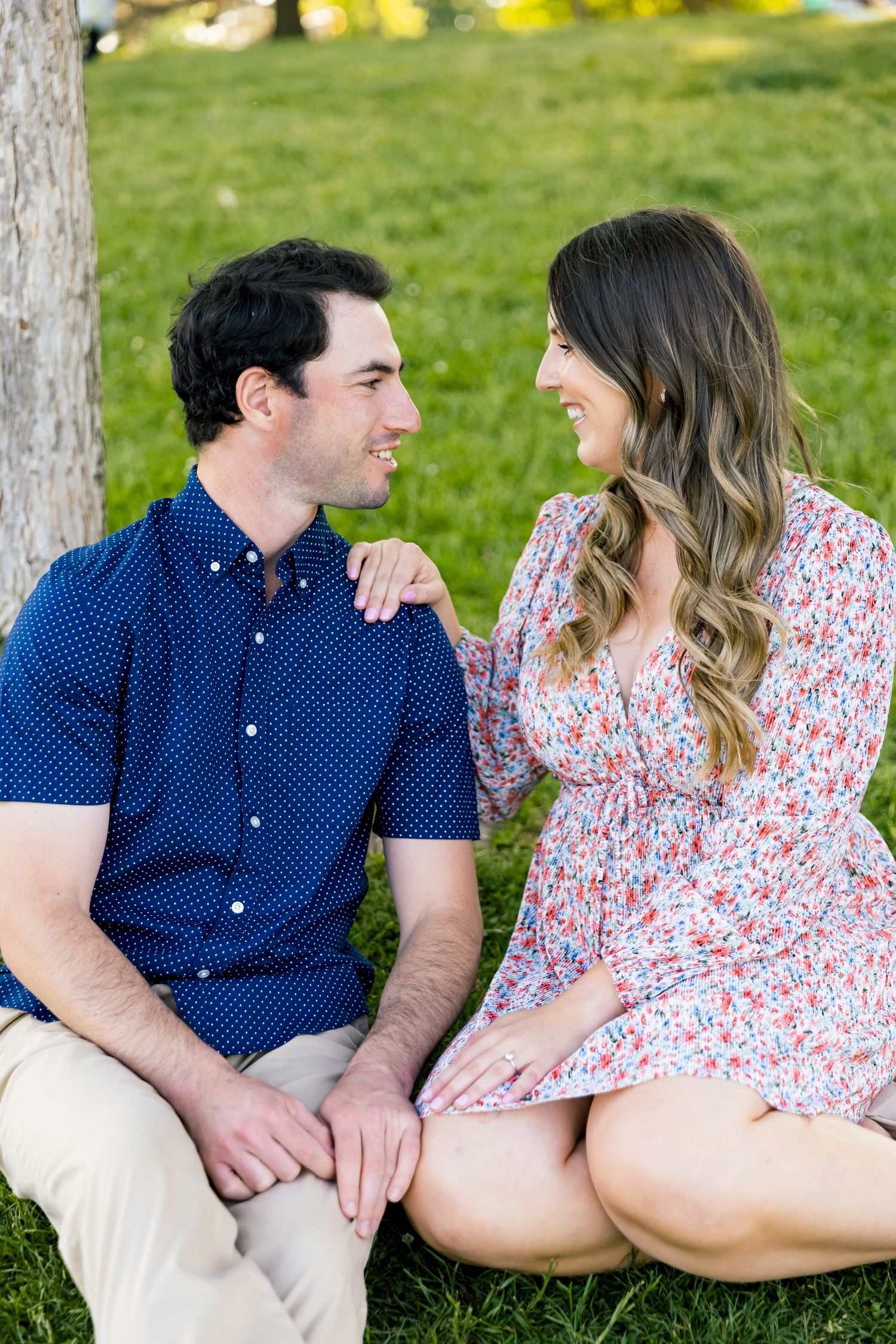 A couple sits close together on grass, sharing a loving moment near a tree.