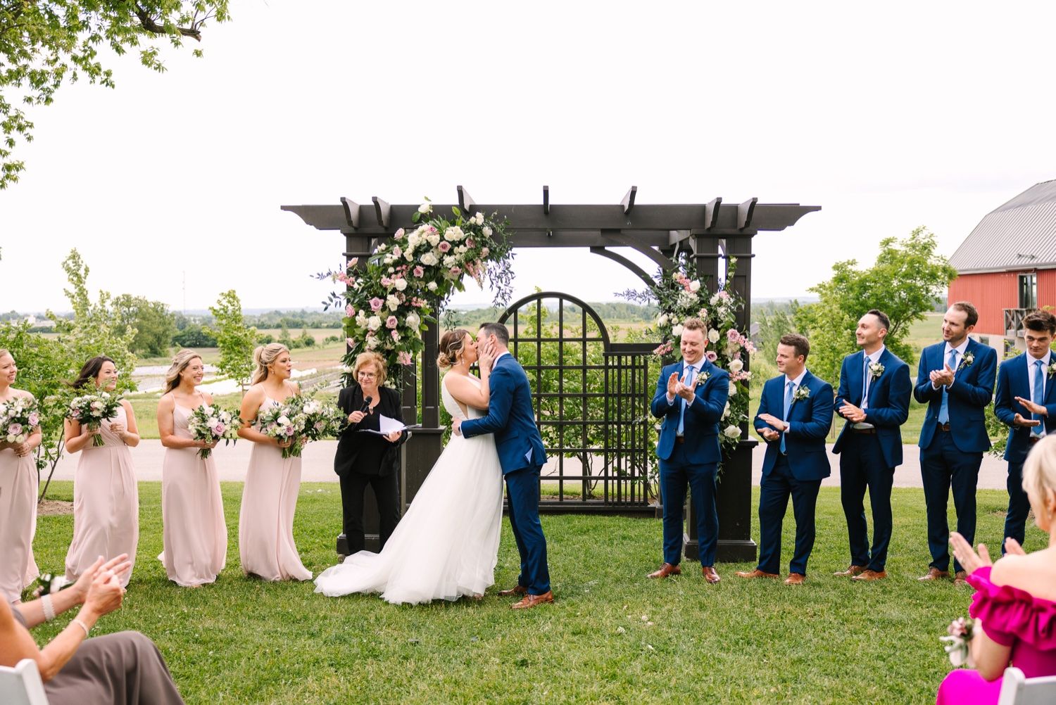 Wedding ceremony at outdoor garden venue with floral arch and bridal party in blue and pink.