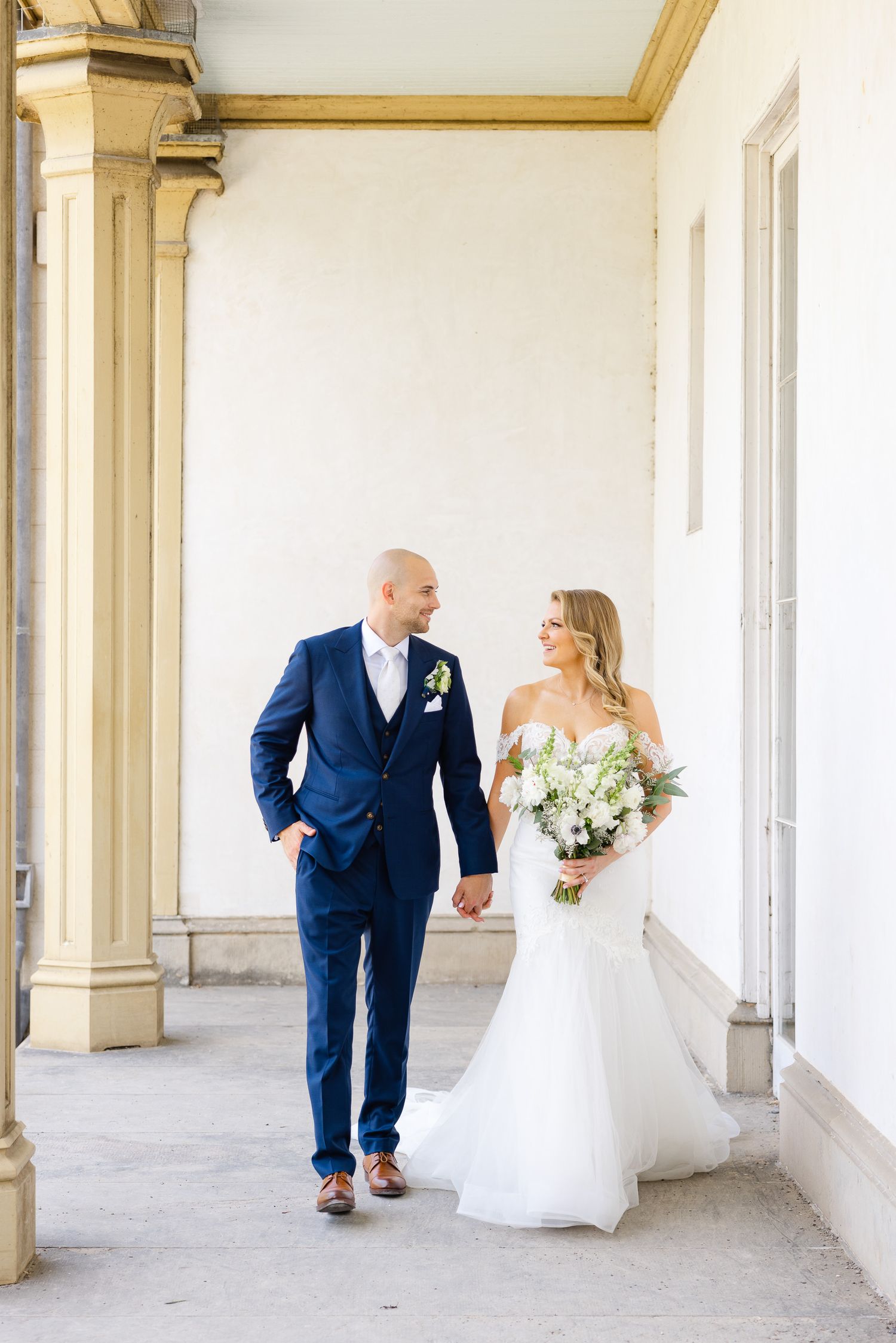 A couple in wedding attire walking along a columned porch on their special day.