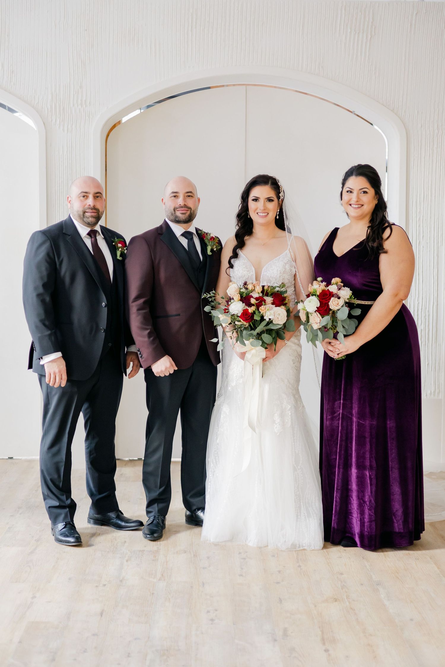 Wedding party poses under white archway with burgundy and cream floral bouquets.