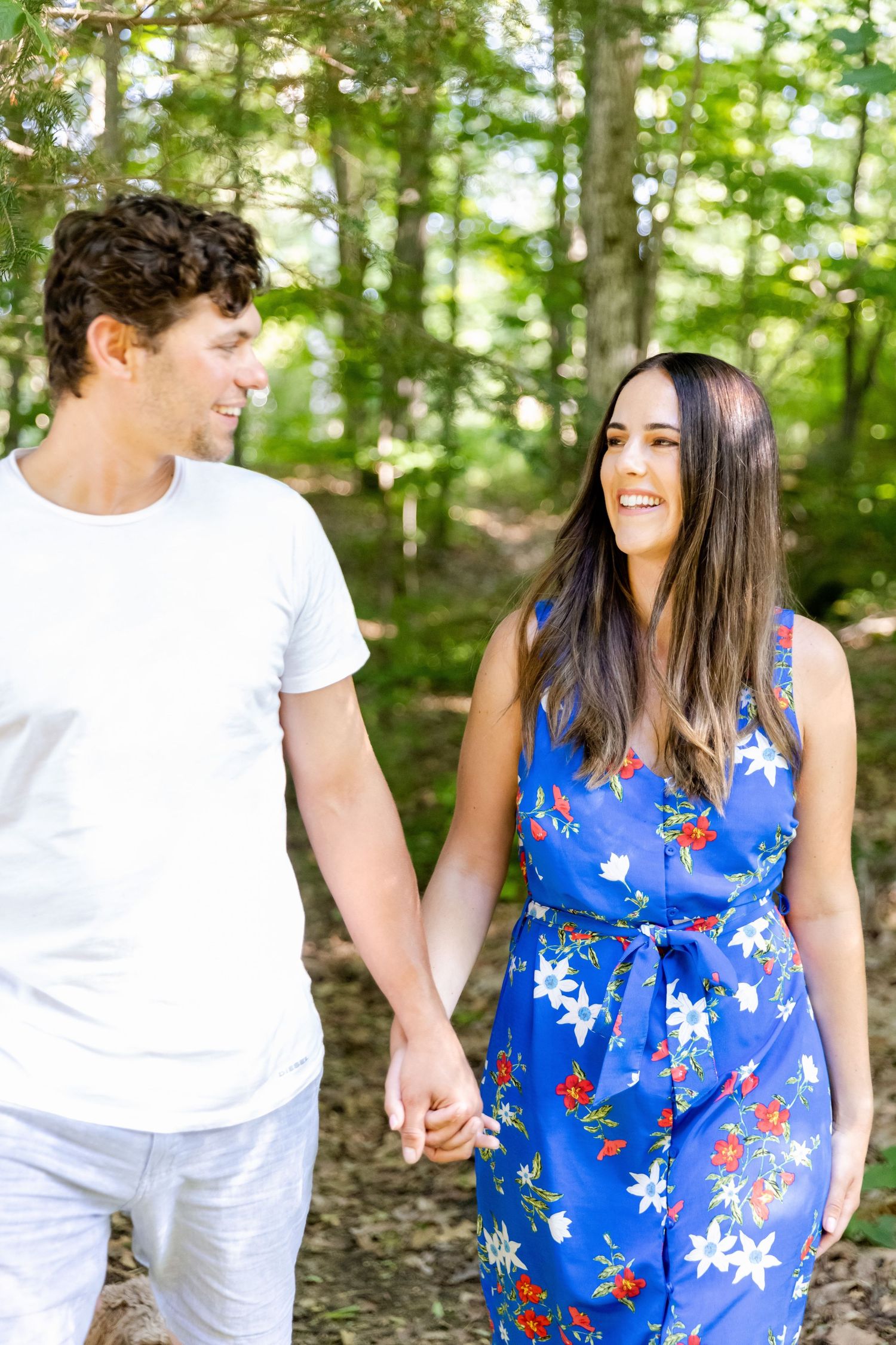 Couple holding hands and exchanging smiles while walking through a sunny forest path.