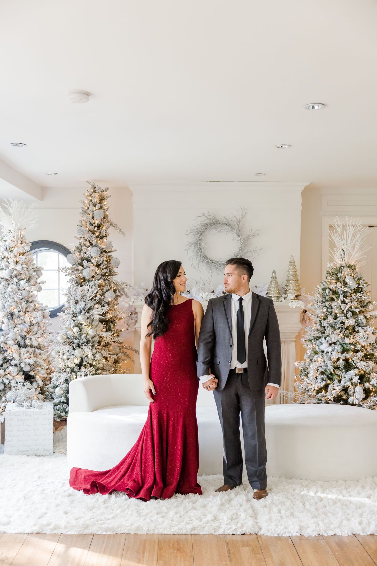 Stunning holiday portrait with red gown and grey suit against white Christmas trees.