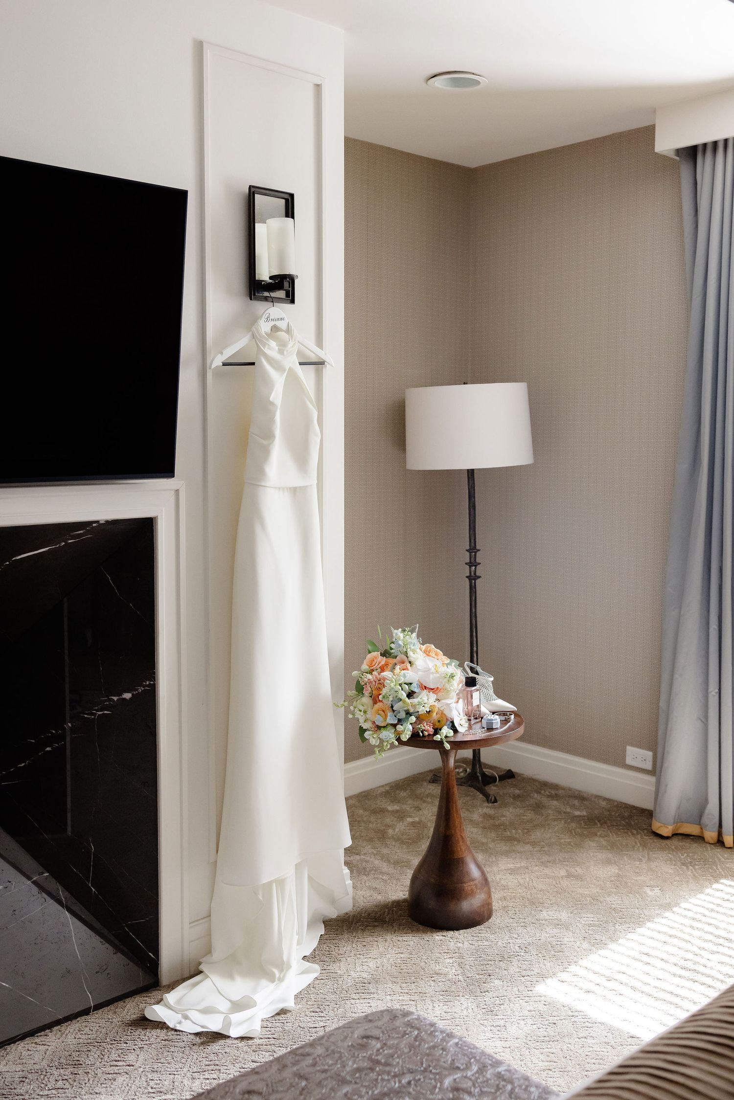 White wedding dress hanging in a modern hotel room with gray walls and black fireplace.