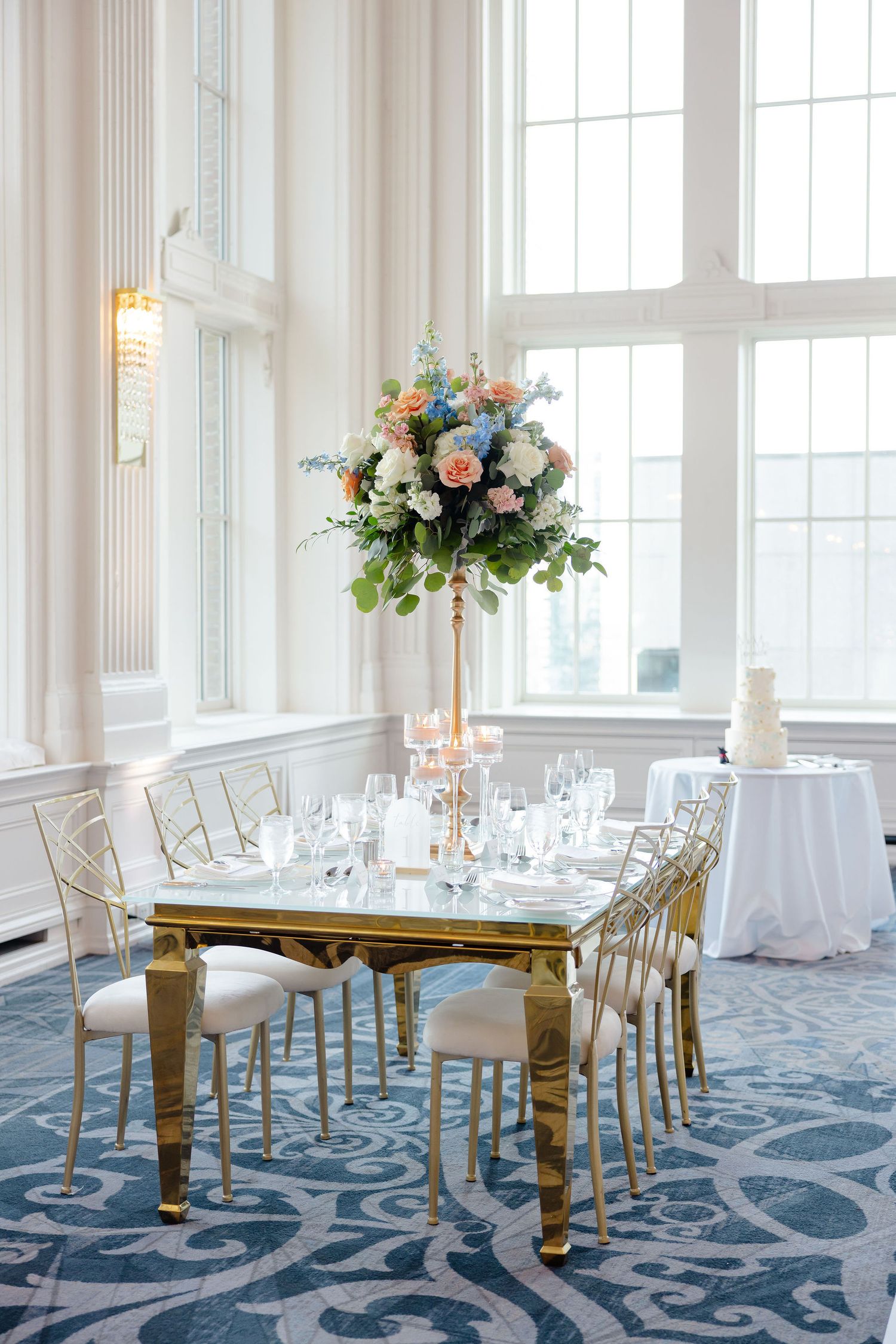 Luxurious wedding reception table with tall floral centerpiece, gold chairs and blue patterned carpet in bright venue.