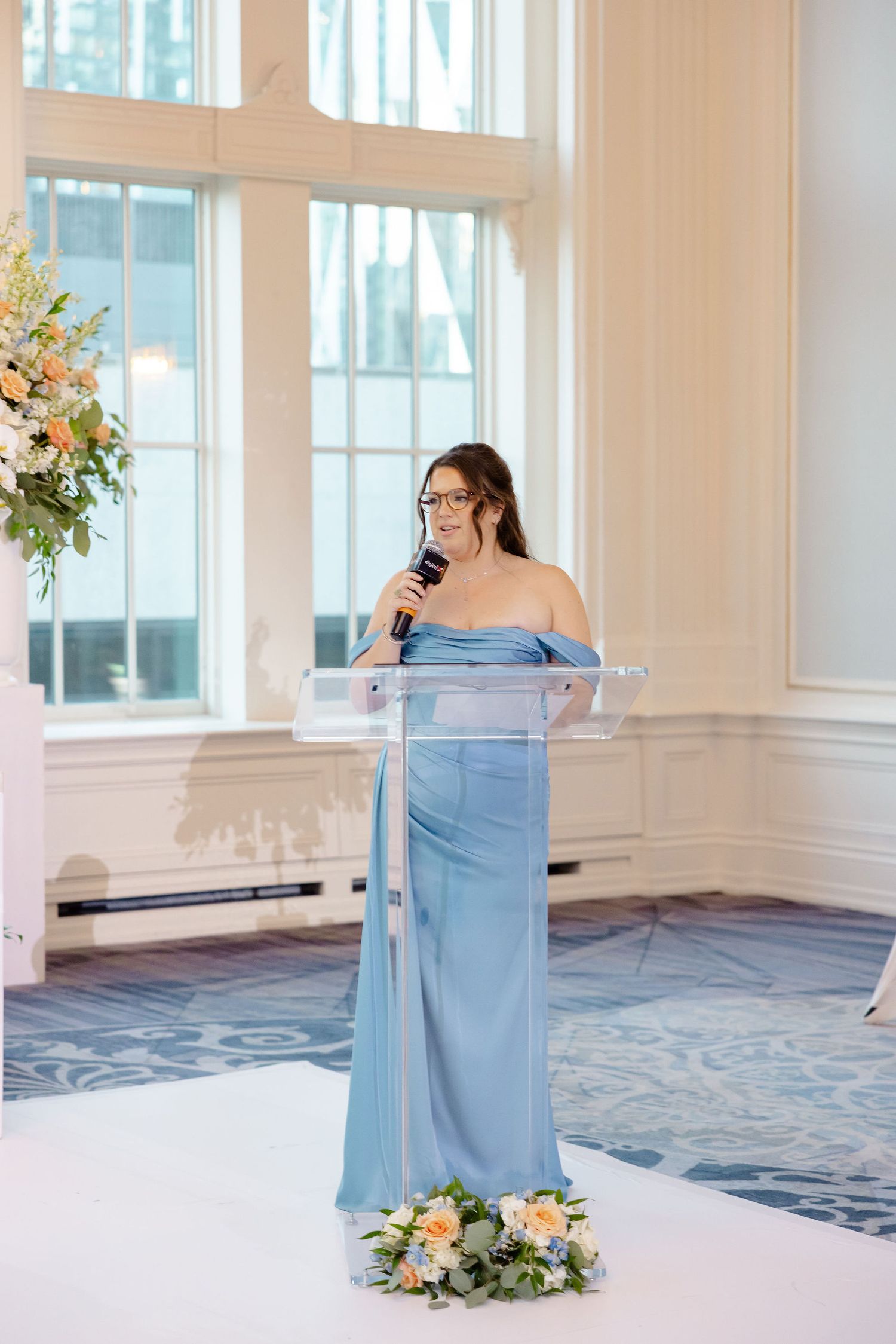 A speaker in a light blue gown delivers remarks at an elegant wedding reception podium.