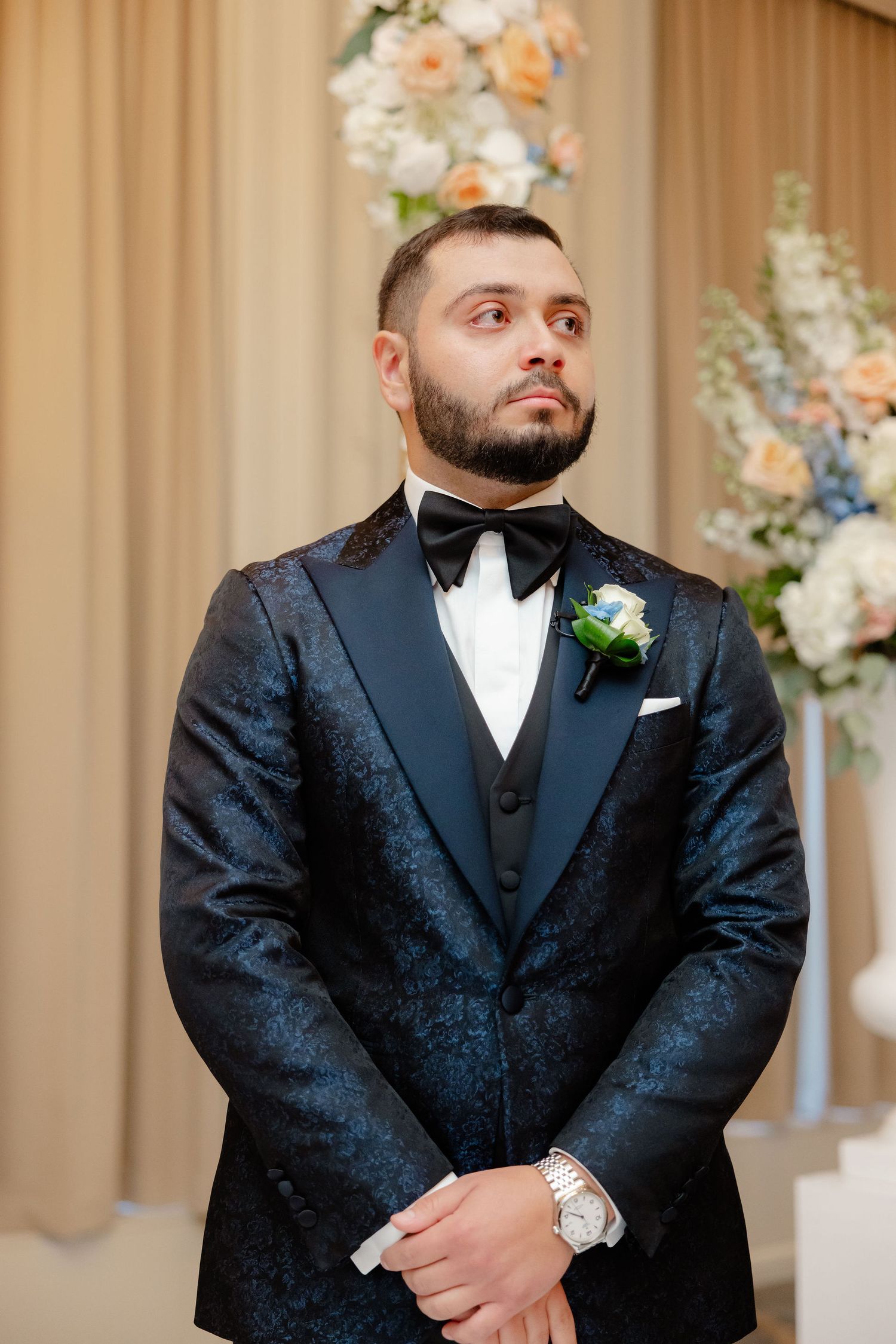 Groom in black textured tuxedo and bowtie waiting at wedding ceremony.