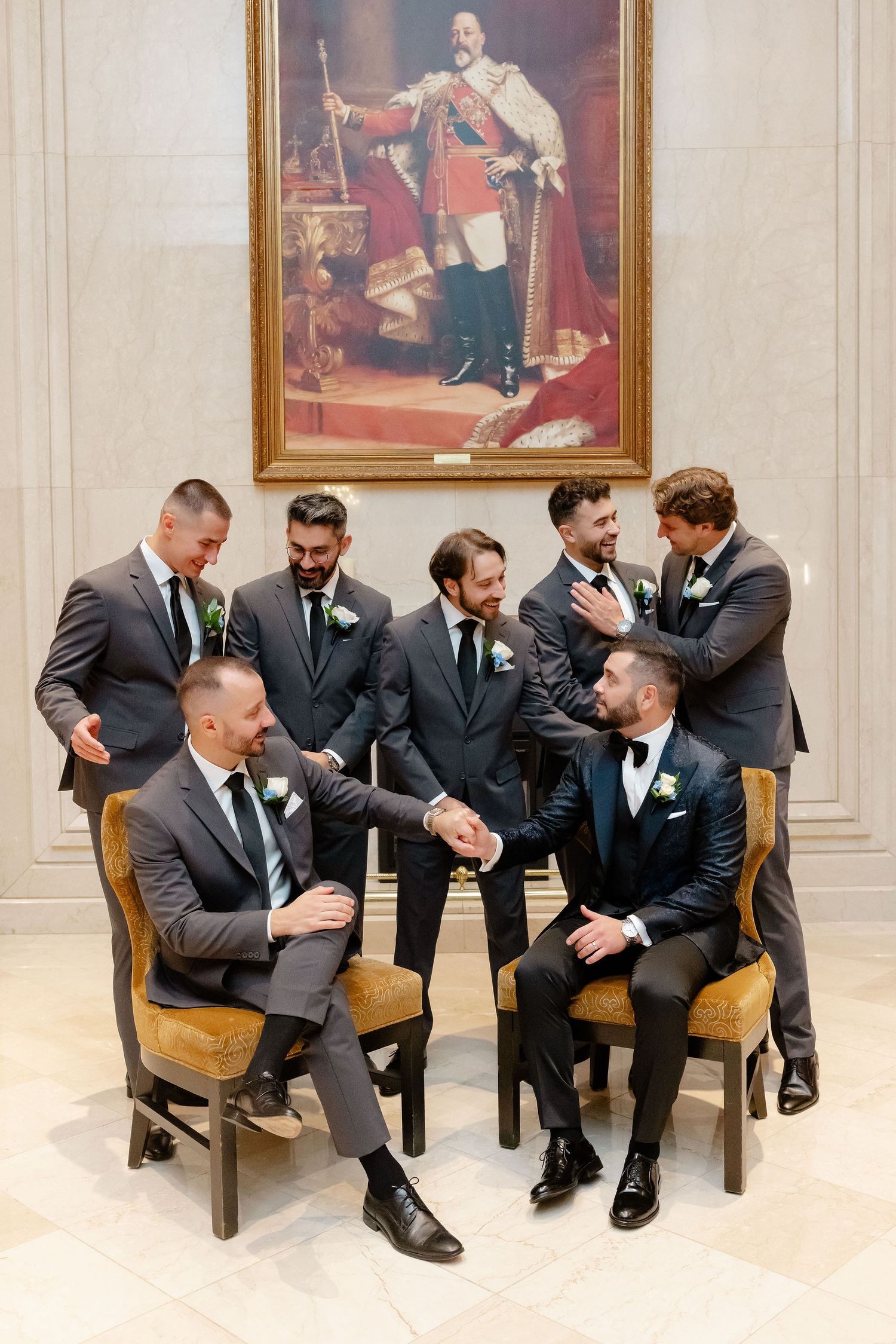 Groomsmen in dark suits share candid laughs while seated on wooden chairs in an elegant room.