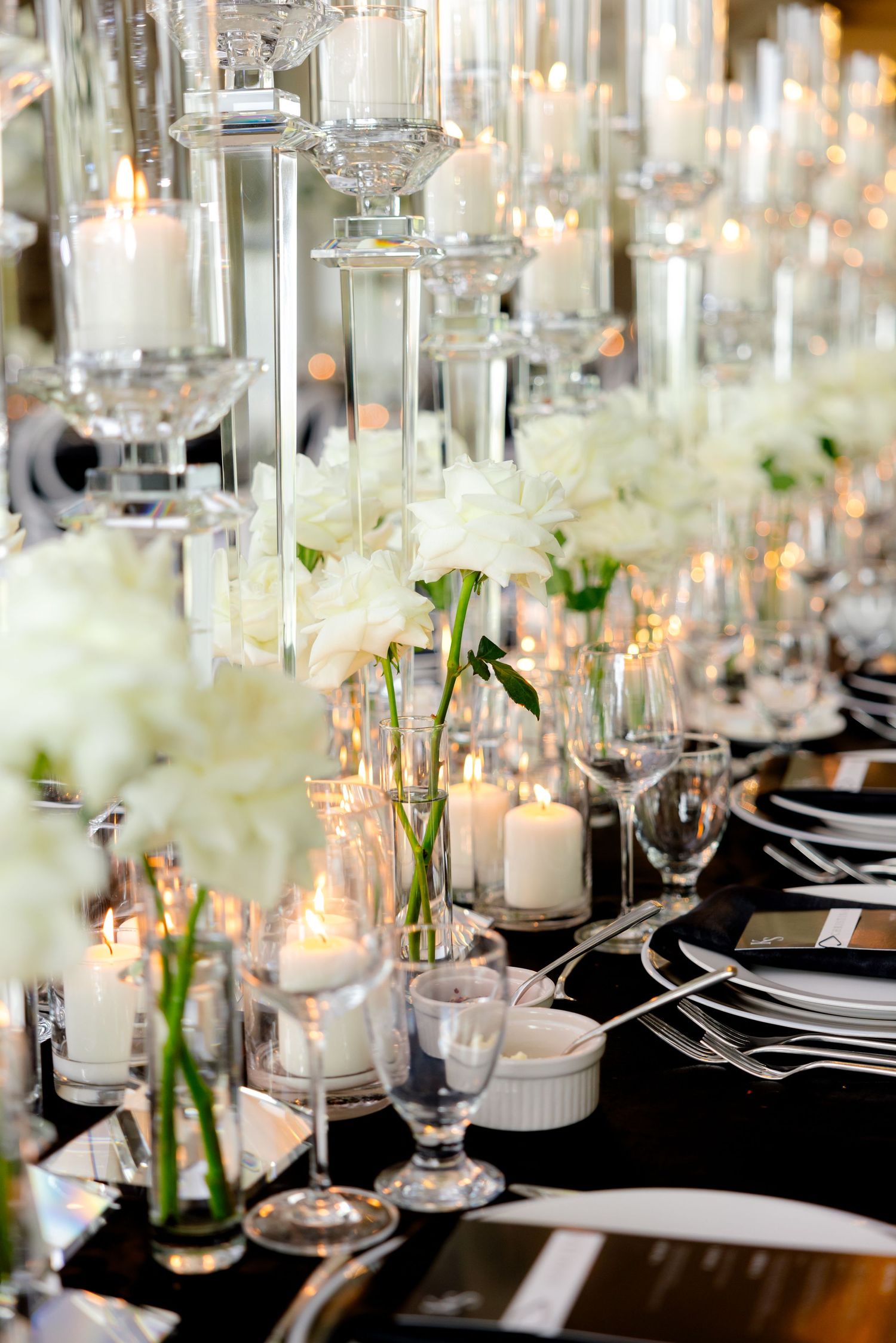 Detailed shot of wedding table decor featuring glass candleholders, white flowers, and place settings on black linens.
