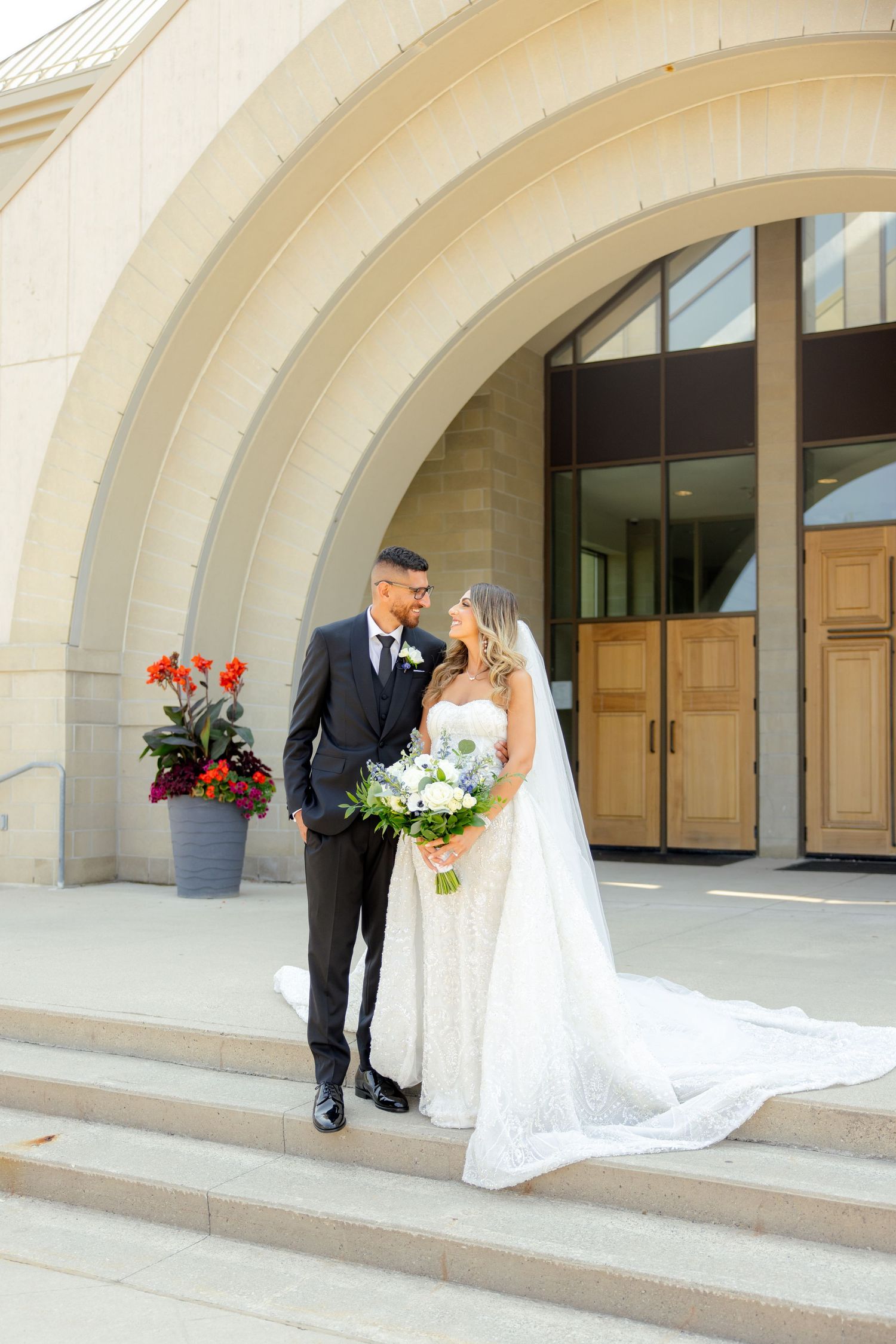 Elegant couple stands on steps of modern building with curved archways and red flowers.