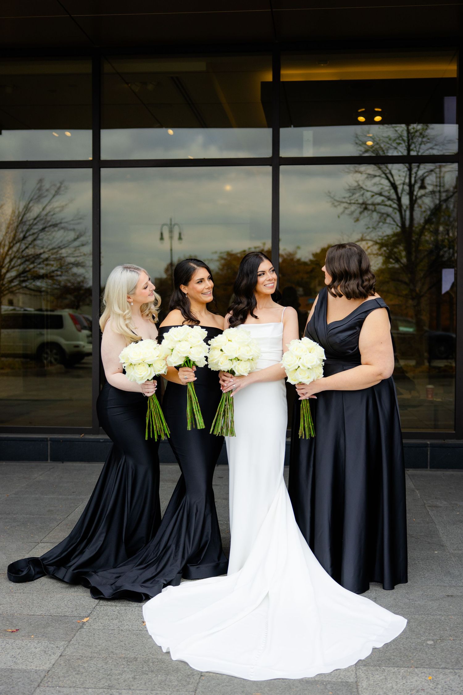 Bridal party in elegant black gowns and white wedding dress pose with cream bouquets against glass building facade.