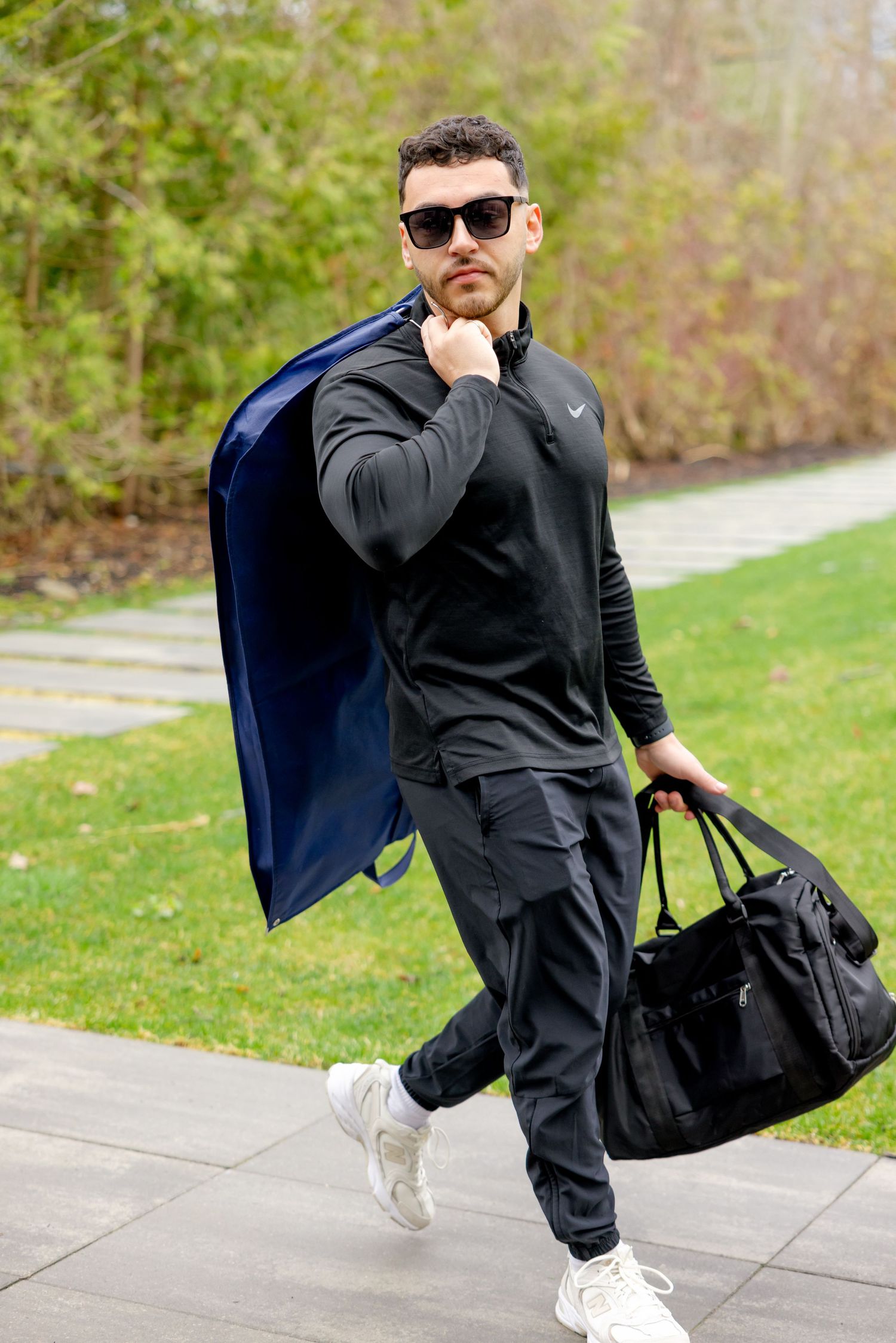 Person in dark sportswear walking outdoors with blue coat and black duffle bag.