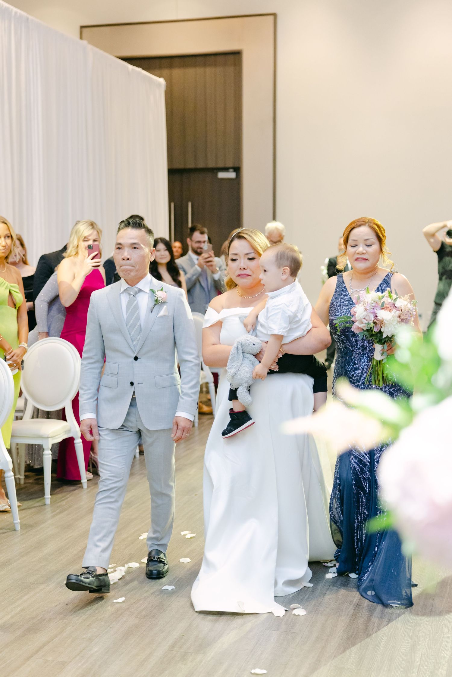 Wedding party members walking down aisle with baby in formal attire at indoor ceremony.