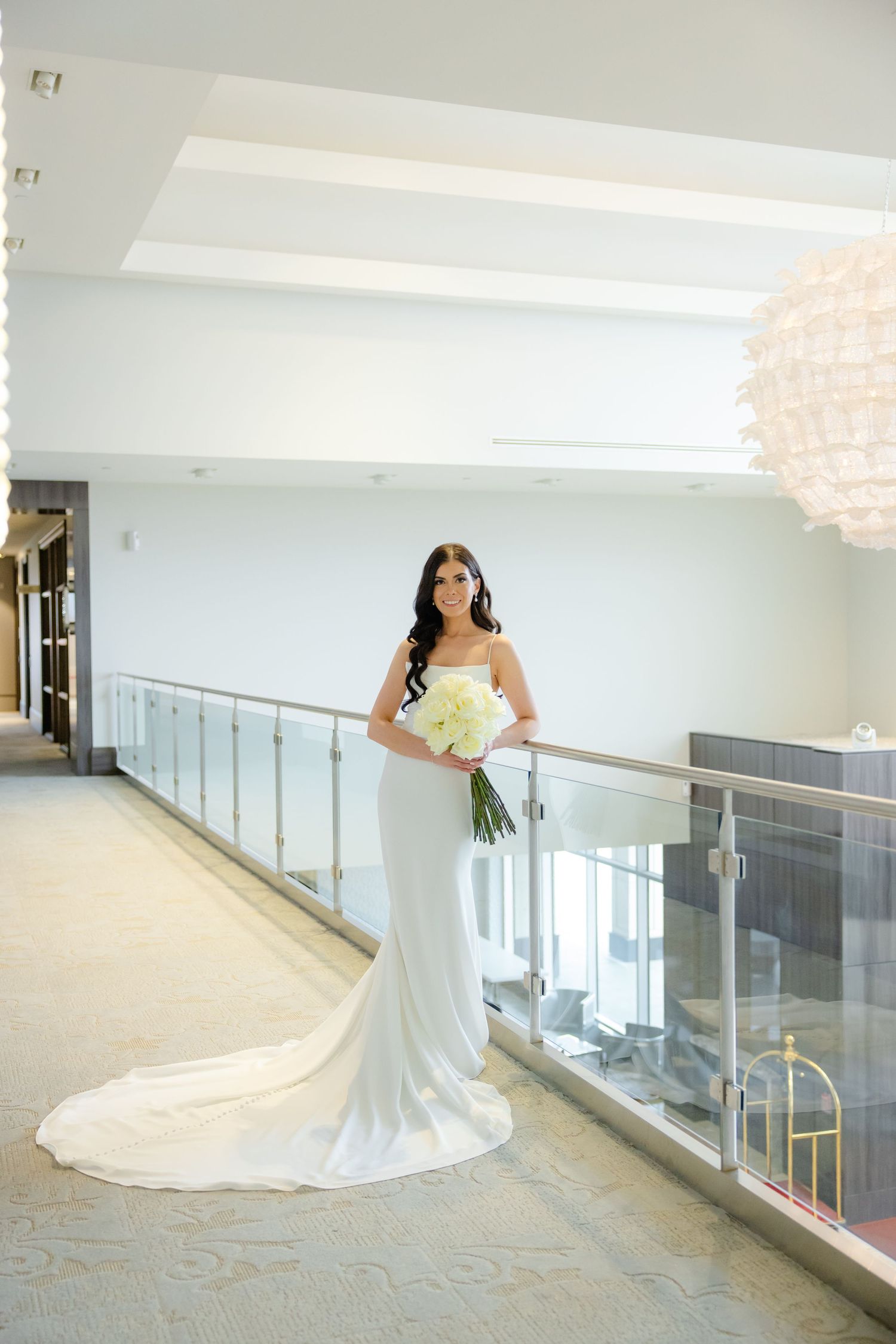 Bride in fitted white gown with train poses alone in modern hallway with glass railings.