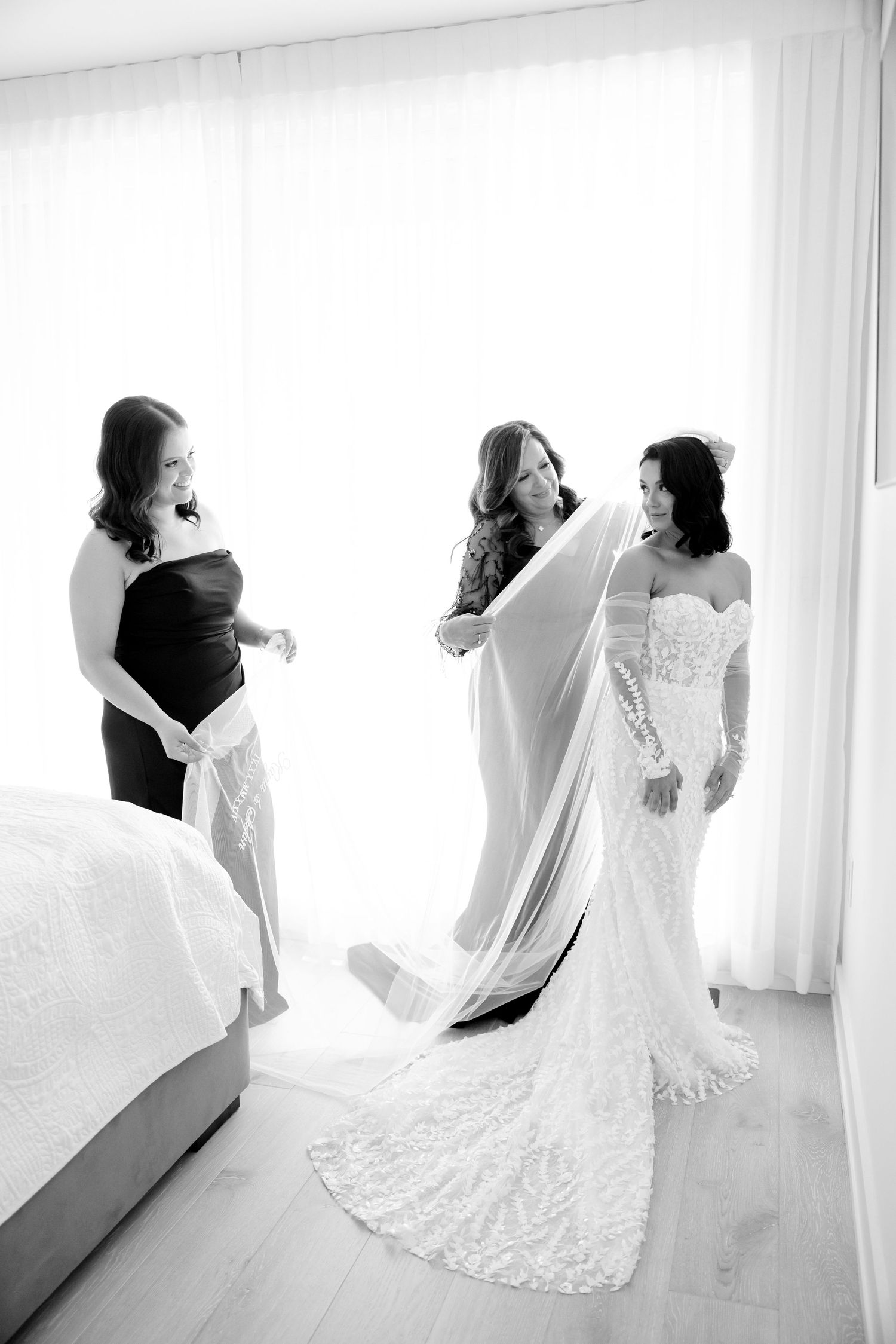 Black and white photo of wedding preparations as bridesmaids help with the bride's veil and gown.