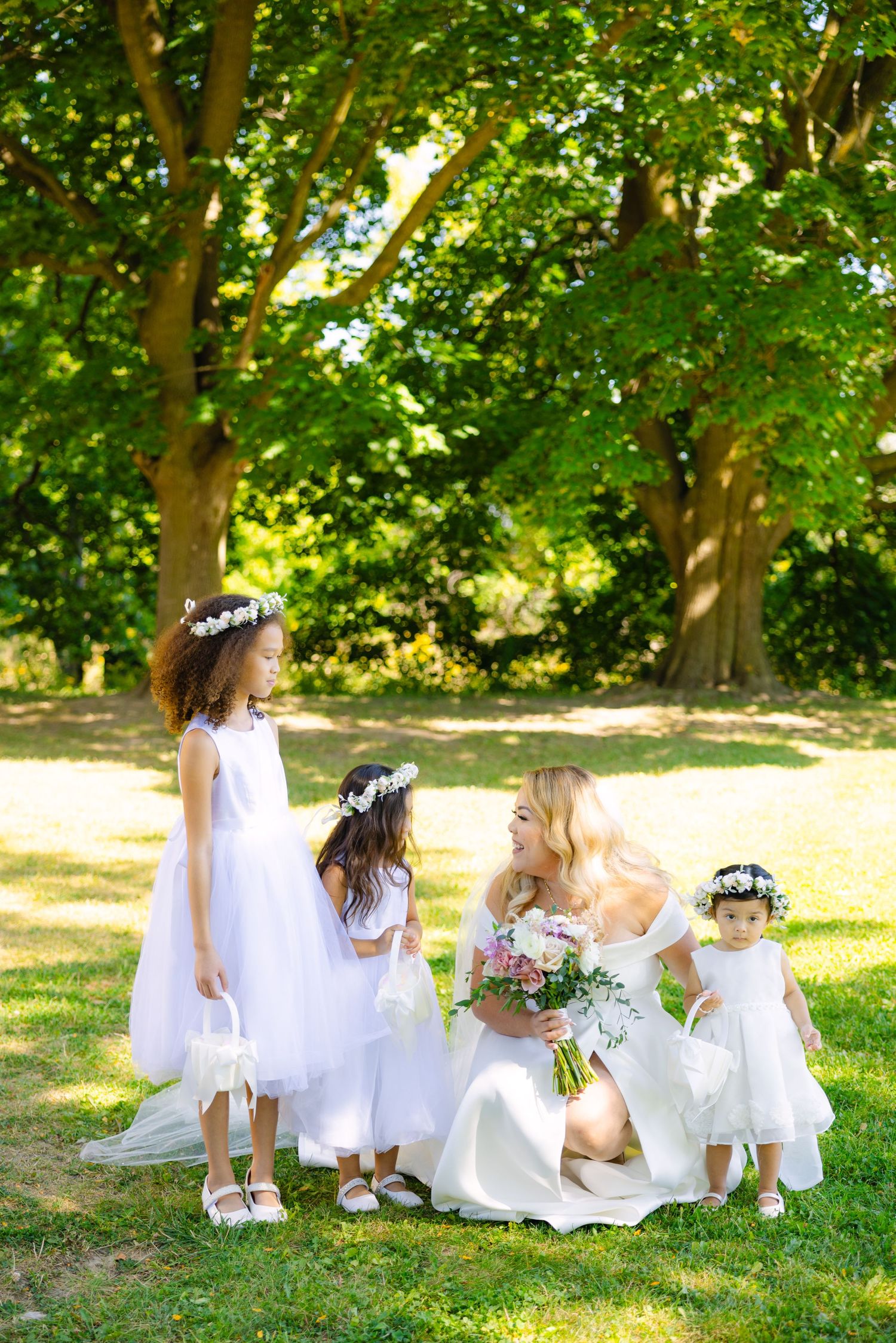 Sweet moment with flower girls in white dresses gathered around bride in garden setting.