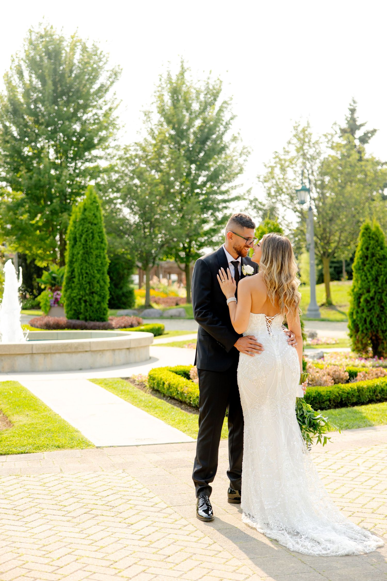 Romantic wedding couple embracing on a brick pathway surrounded by perfectly manicured gardens and trees.