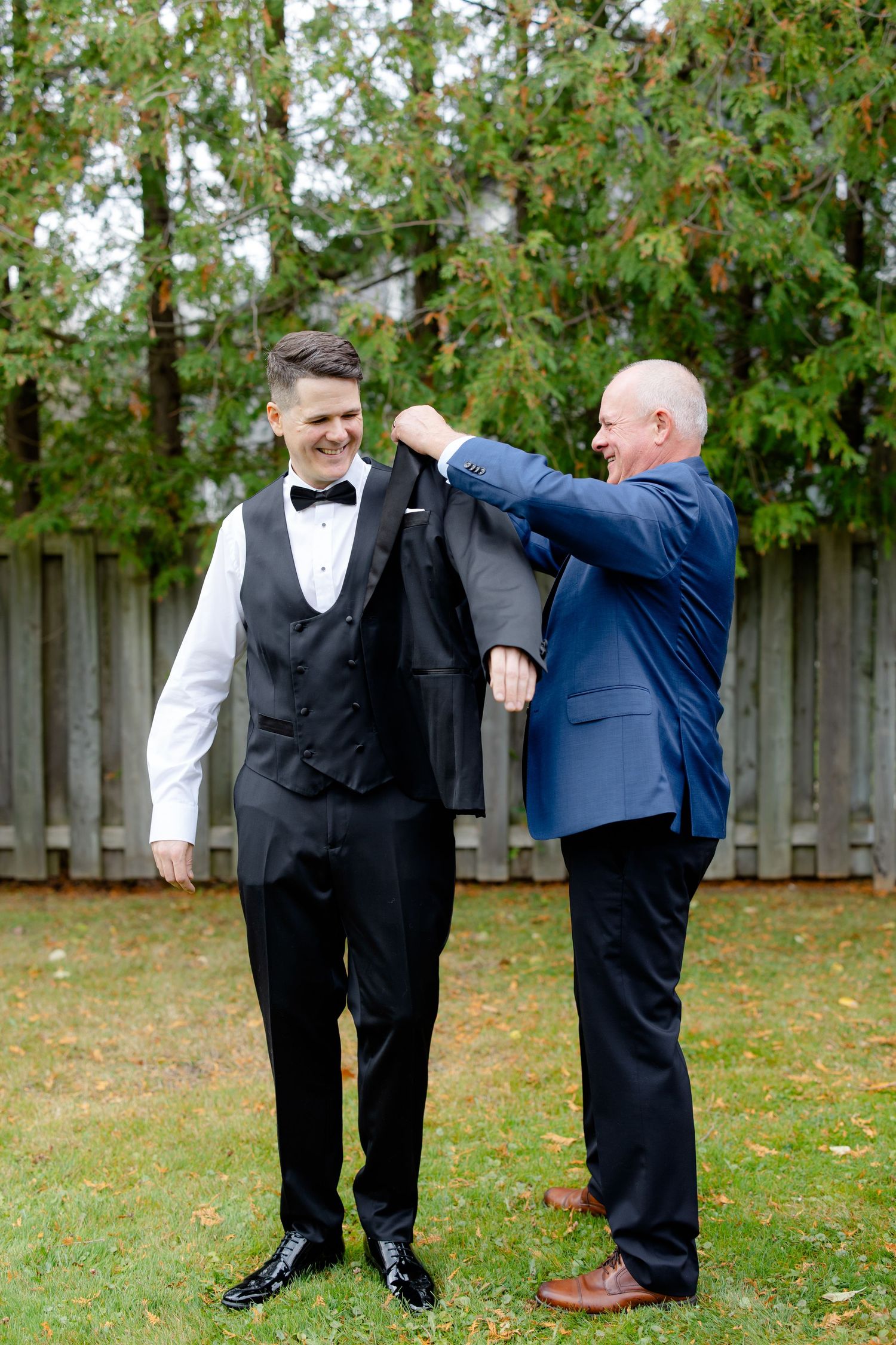 A groom gets help putting on his formal black suit jacket in an outdoor garden setting.