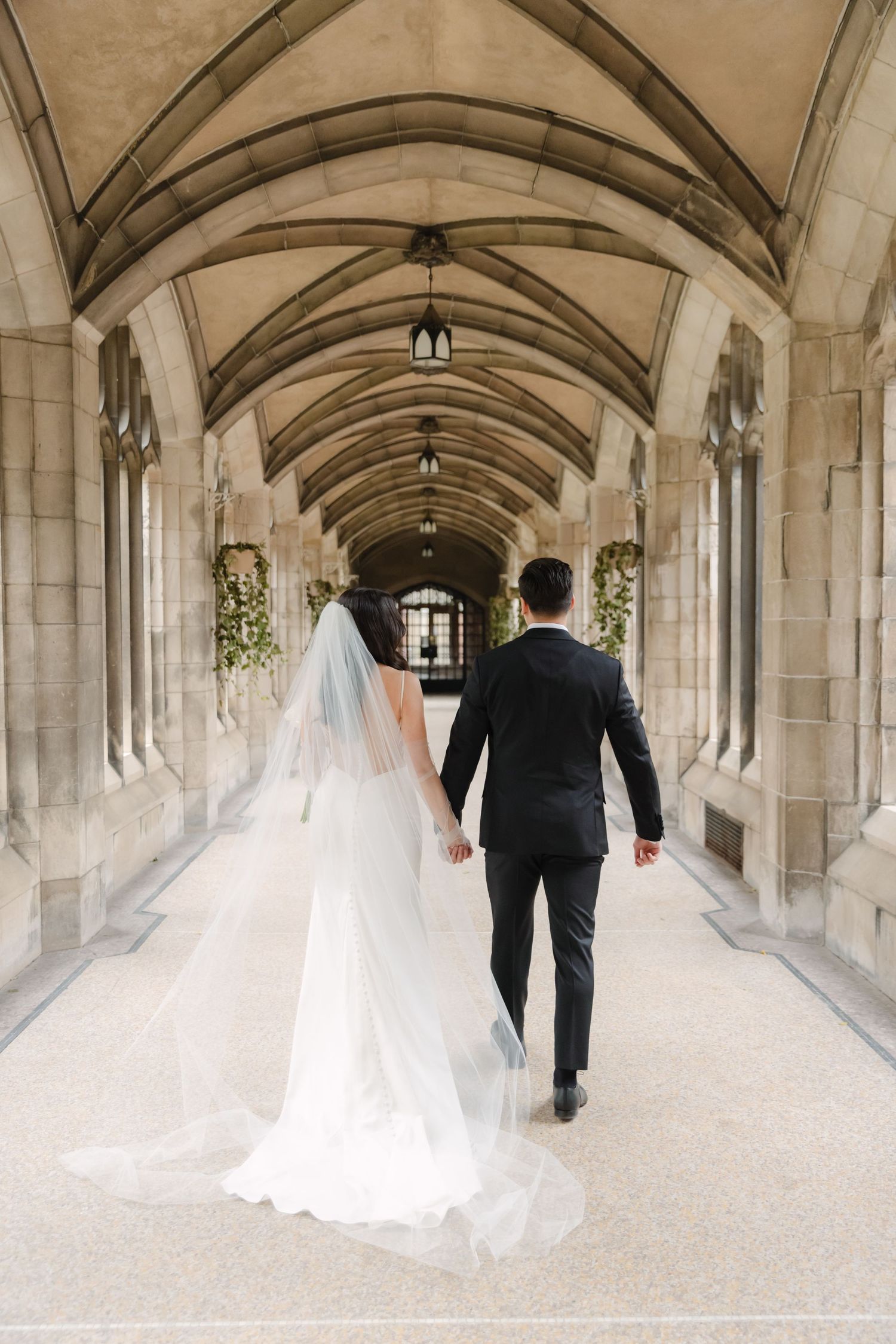 Newlyweds walk hand in hand down a gothic stone corridor decorated with hanging greenery.