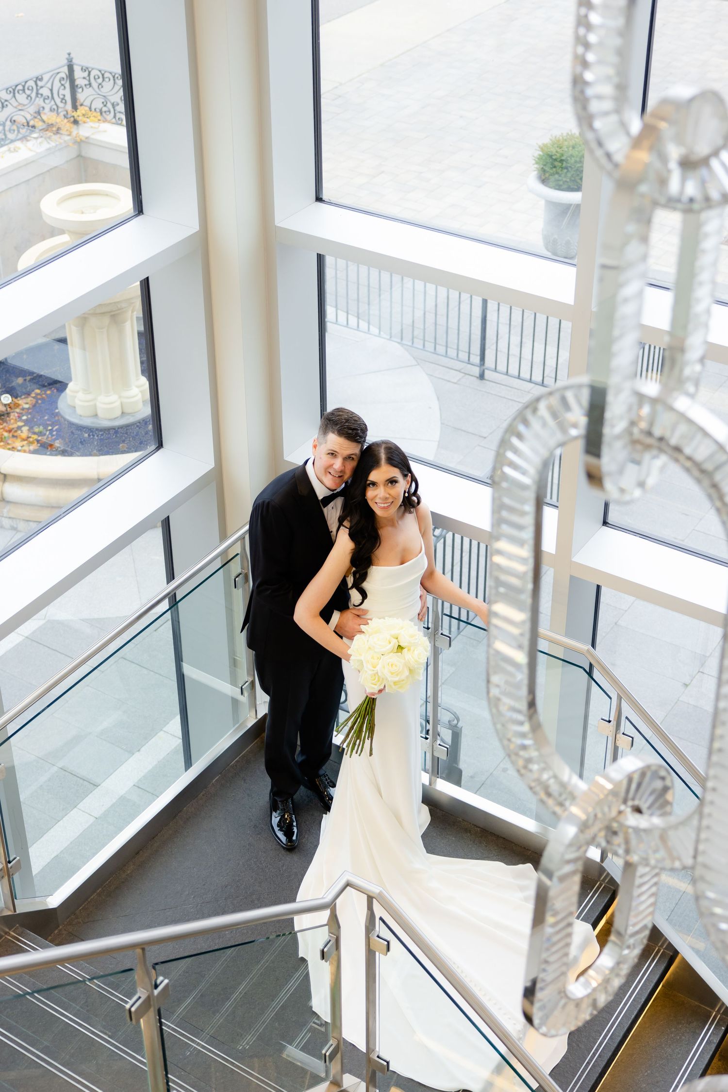 Aerial view of a wedding couple on modern stairs with floor-to-ceiling windows and crystal chandelier details.