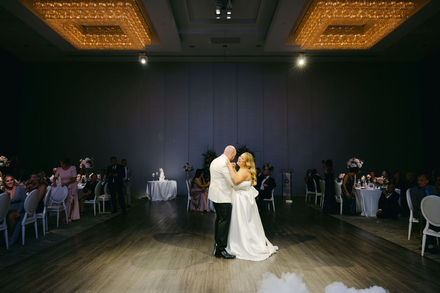 A couple shares their first dance on a dark wooden floor surrounded by reception tables.