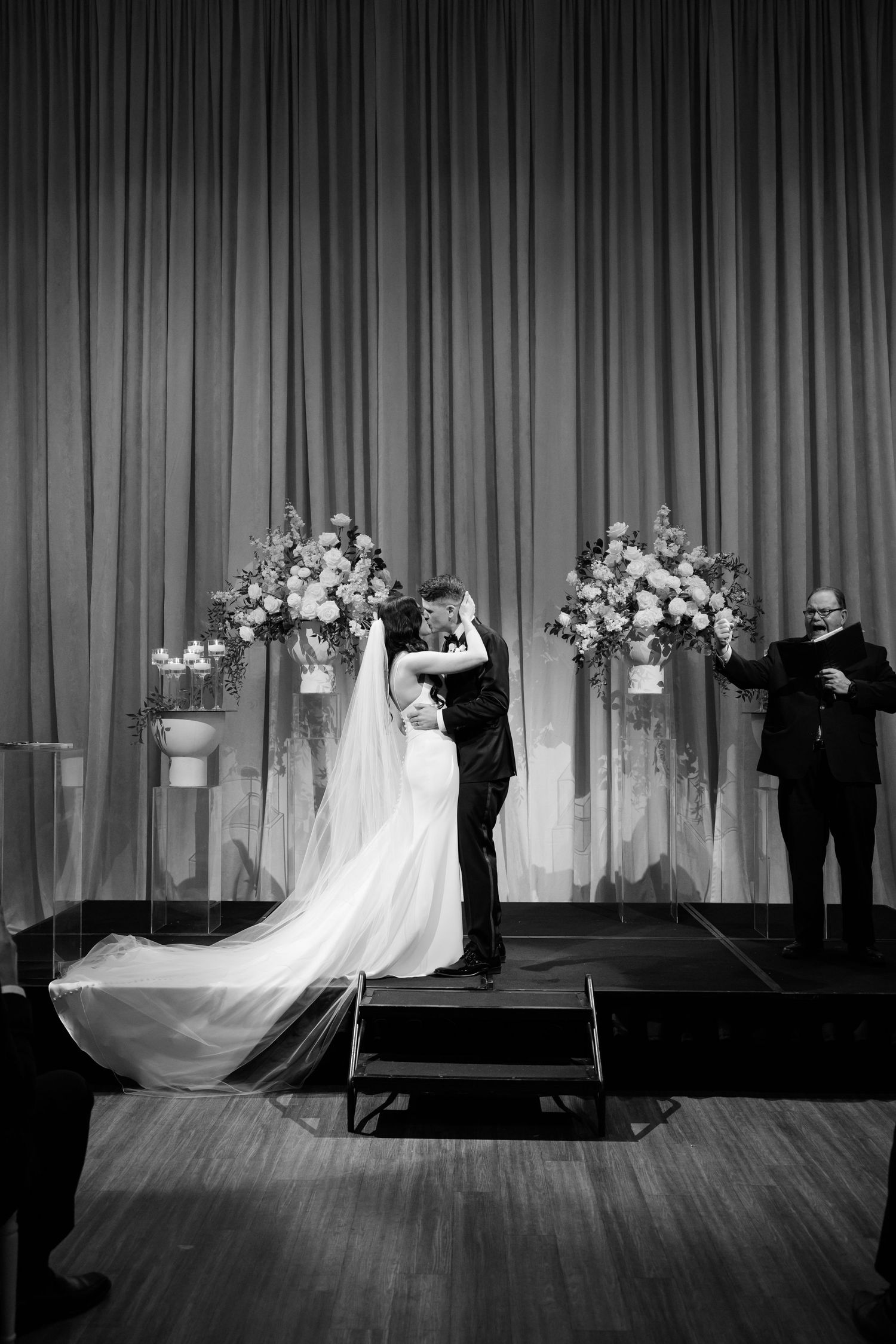 Black and white wedding ceremony photo with couple on stage between tall floral arrangements.